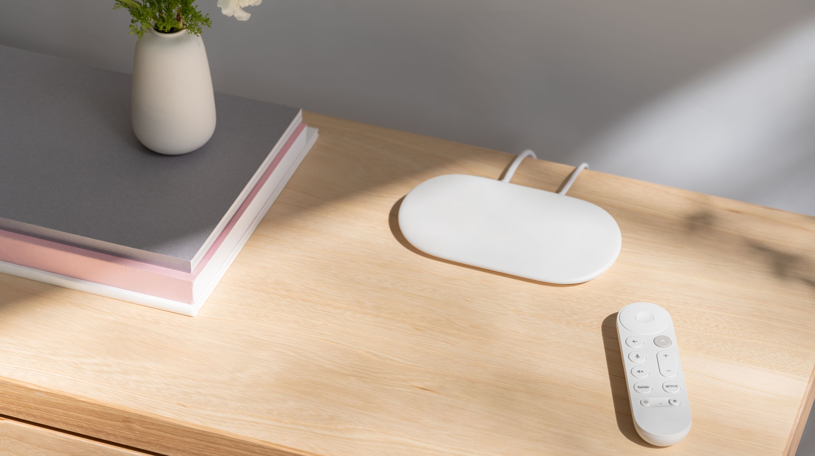 The new Google TV Streamer on a wooden table next to its remote, books, and a plant.