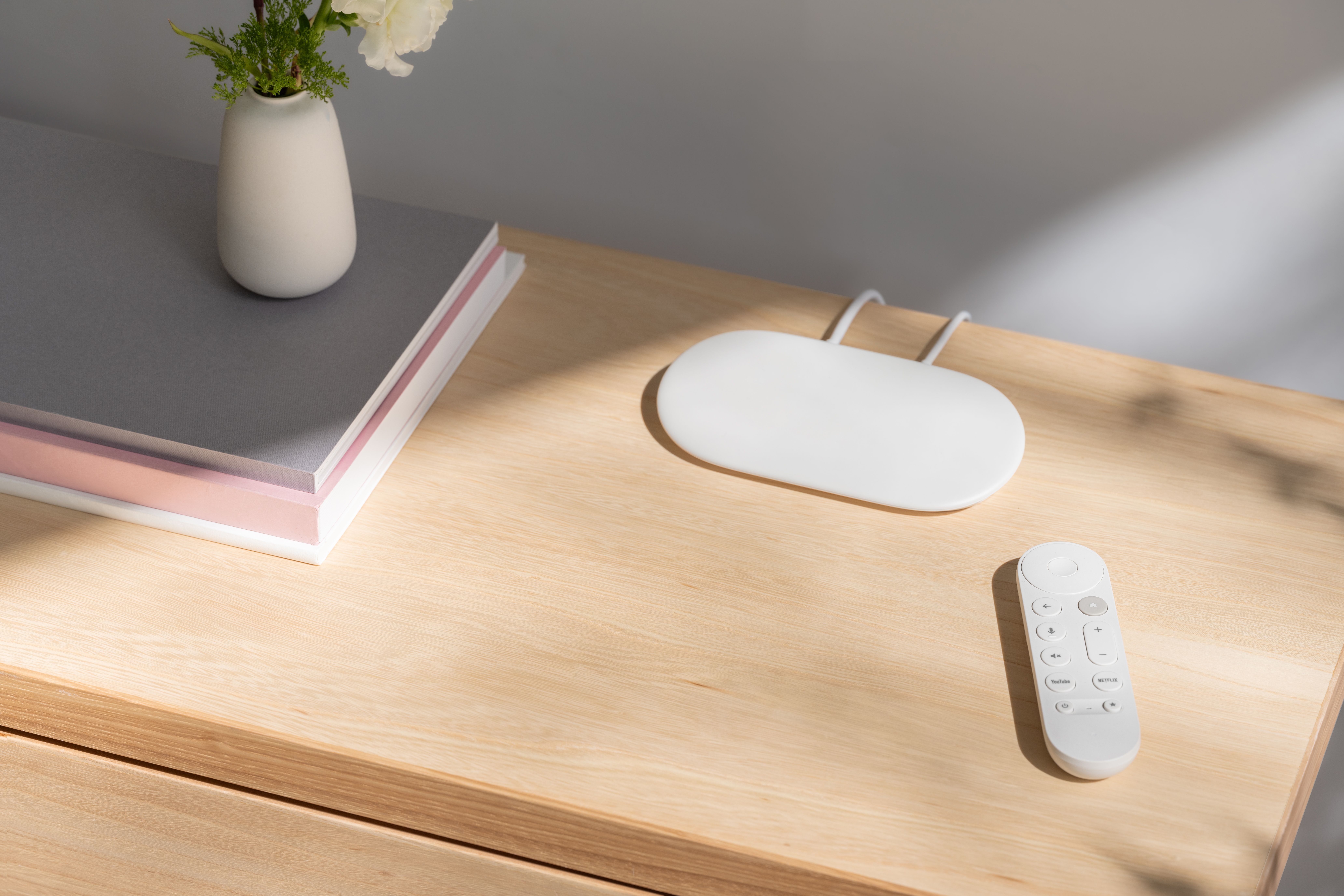 A top view image of the Google TV streamer and its remote on a wooden table next to books and a plant.