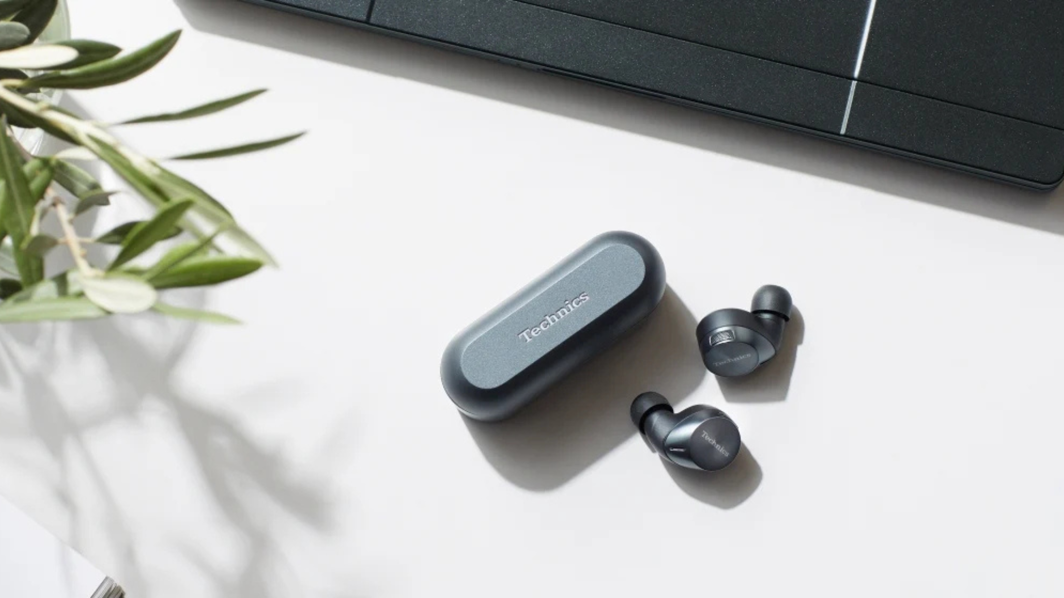 Technics EAH-AZ60 earbuds on a table next to a plant and laptop