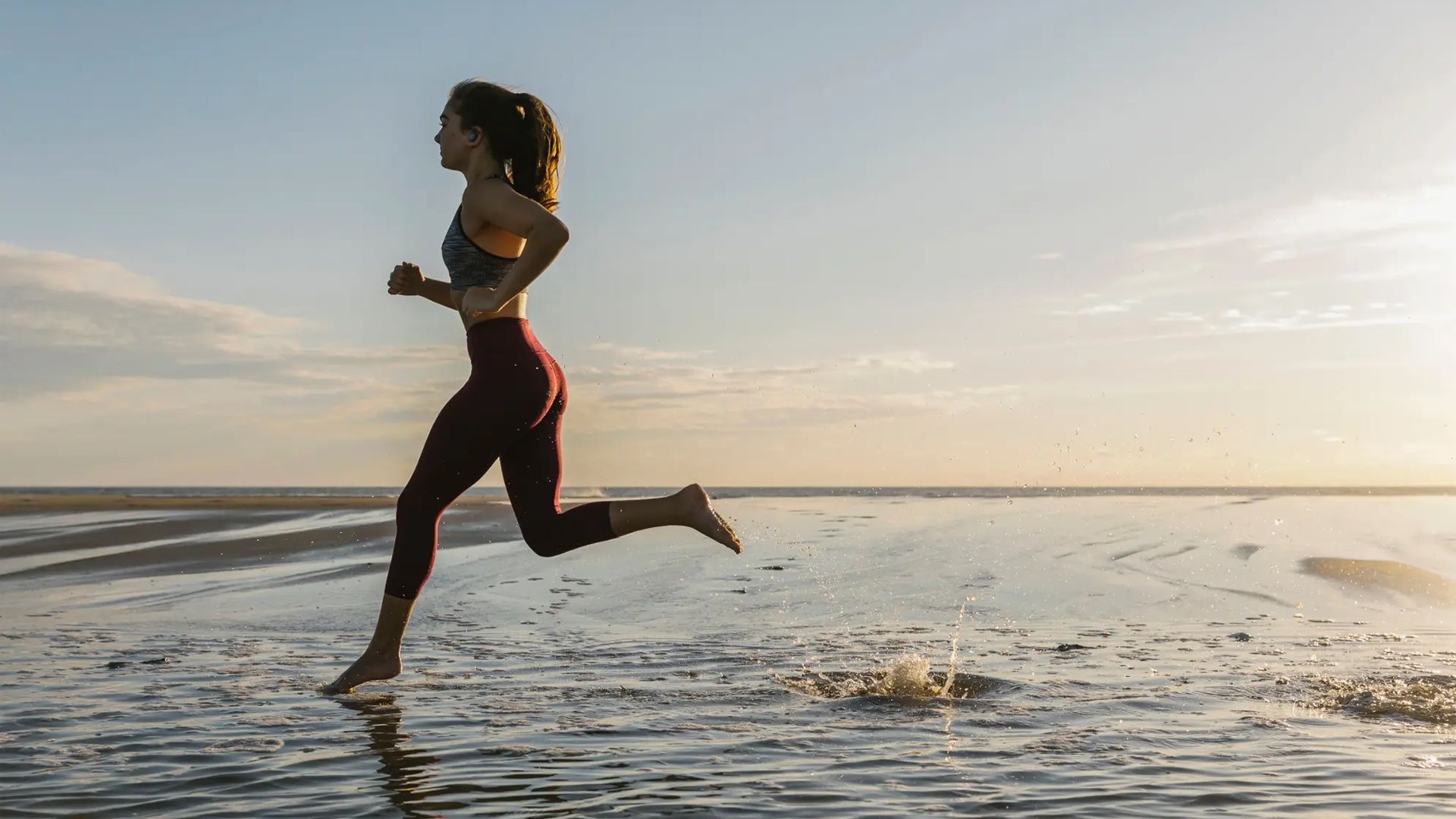 A woman runs along the beach wearing JBL Endurance Race earbuds.