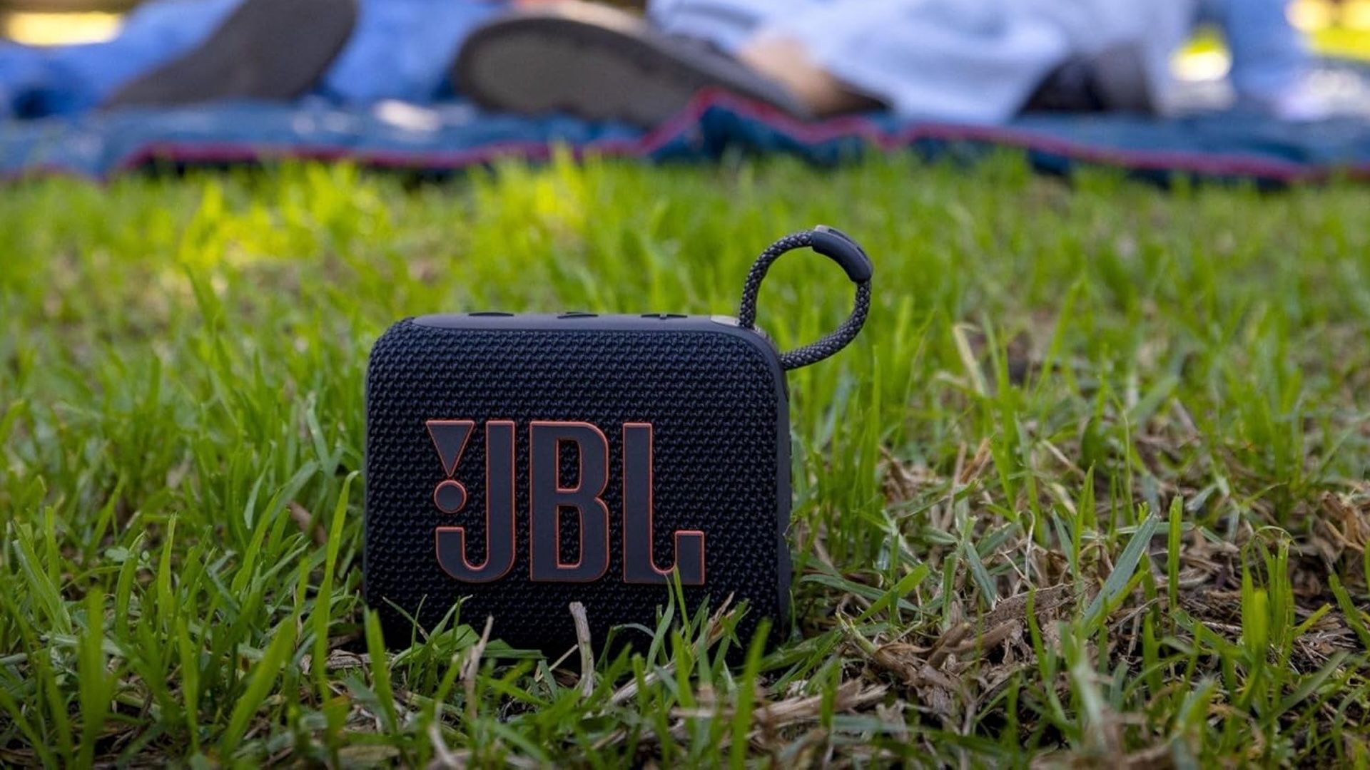 The JBL Go 4 resting in the grass with people at a picnic in the background.