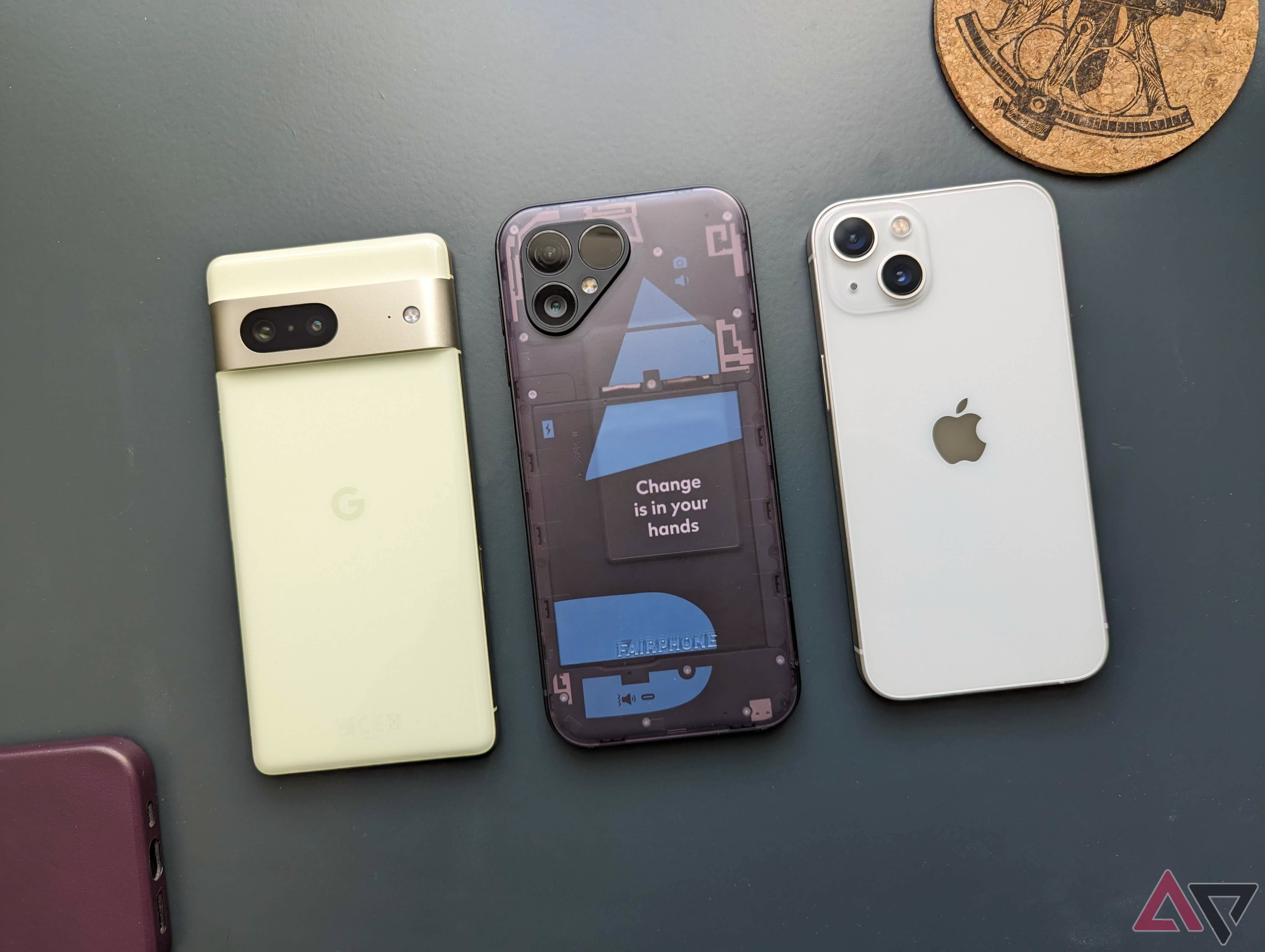 The Google Pixel 7, the Fairphone 5, and the Apple iPhone 13 next to each other on a green background with random items sprinkled around them