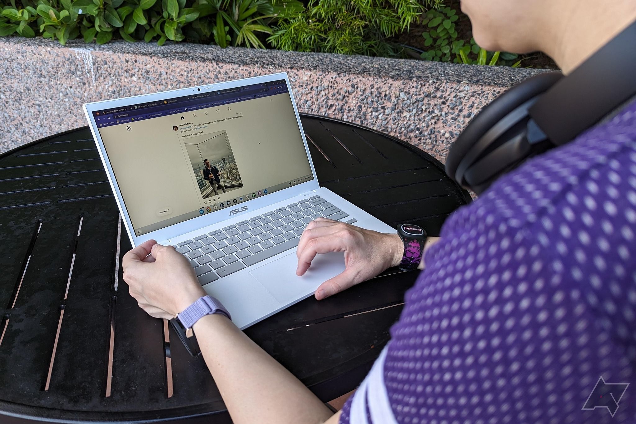 An Asus Chromebook Plus CX34 on a black table outside being used by a person with a purple shirt and headphones on