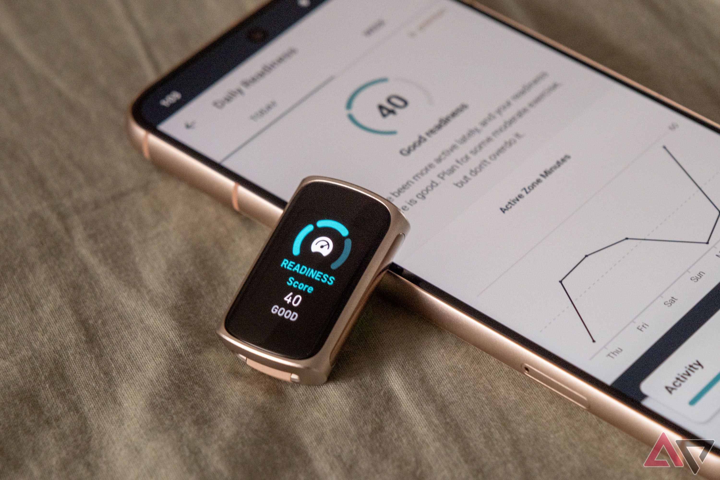 A Fitbit Charge 6 without bands showing a readiness score leaning on a smartphone with the Fitbit app open to the readiness score page on a wooden table.