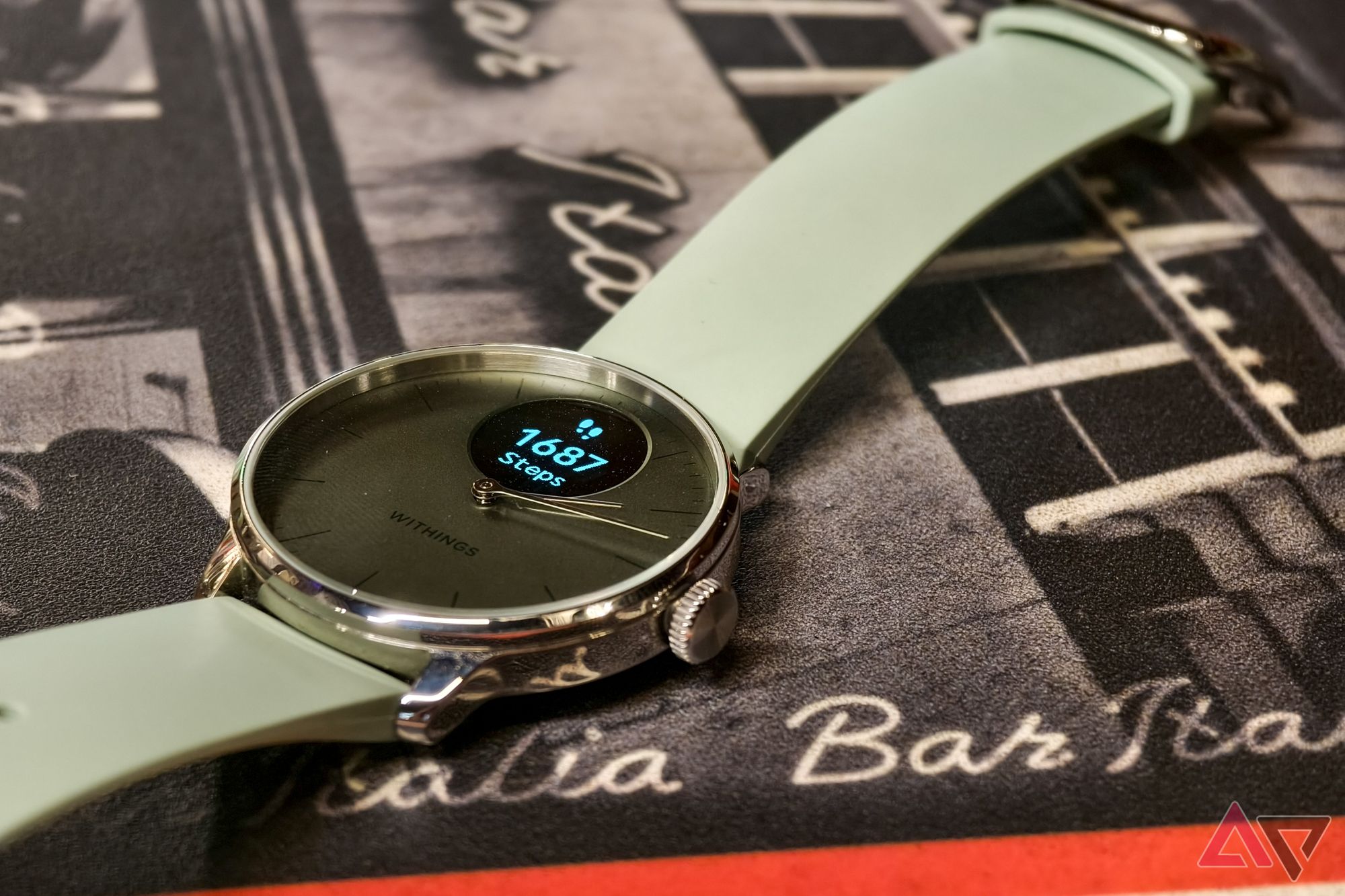 Withings ScanWatch Light in Green, with the display on, photographed from above at an angle with a close-up on the watch face showing step count, resting on an Italian restaurant table