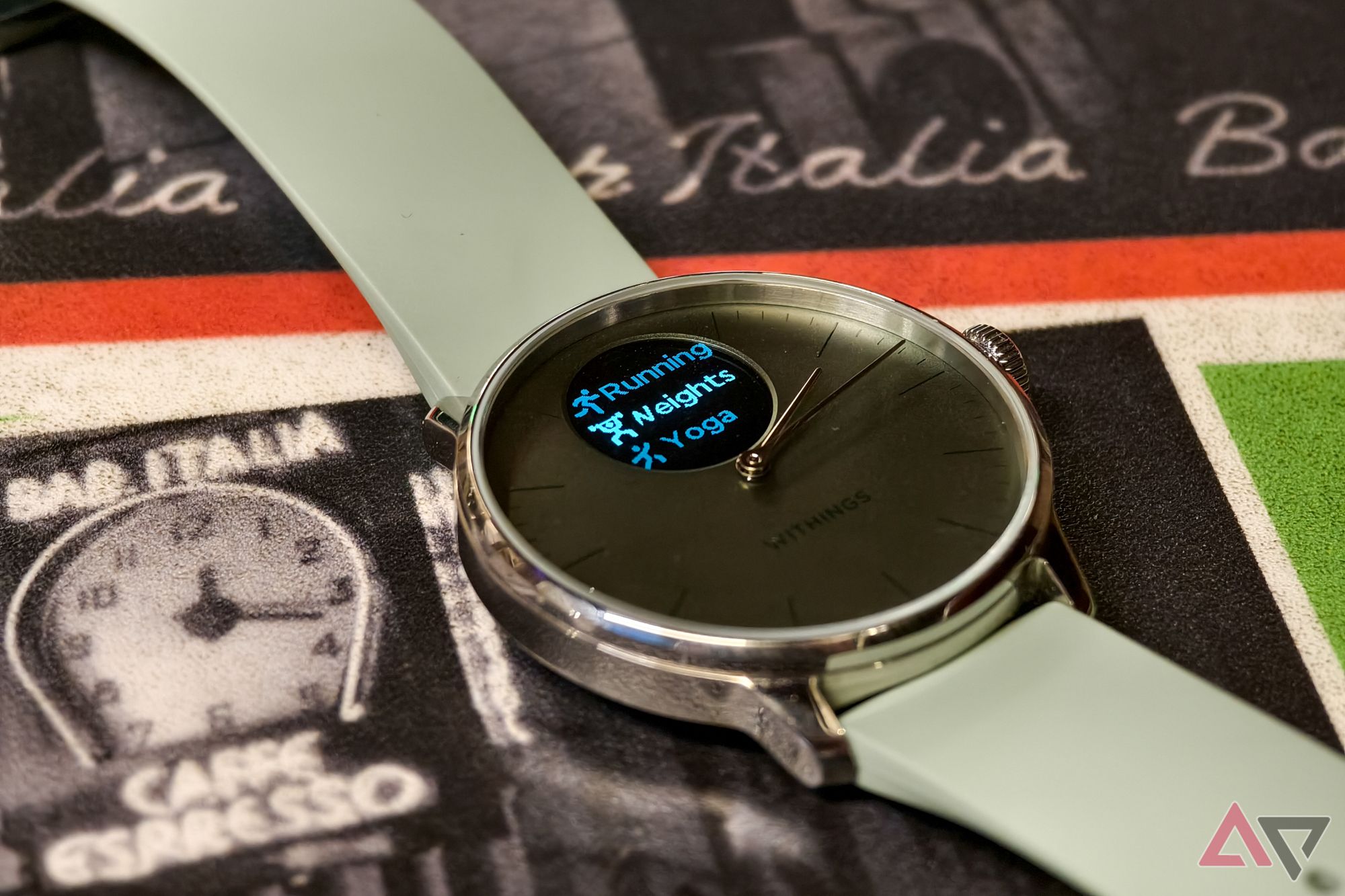 Withings ScanWatch Light in Green, with the display on, photographed from above at an angle with a close-up on the watch face showing a list of workout modes, resting on an Italian restaurant table