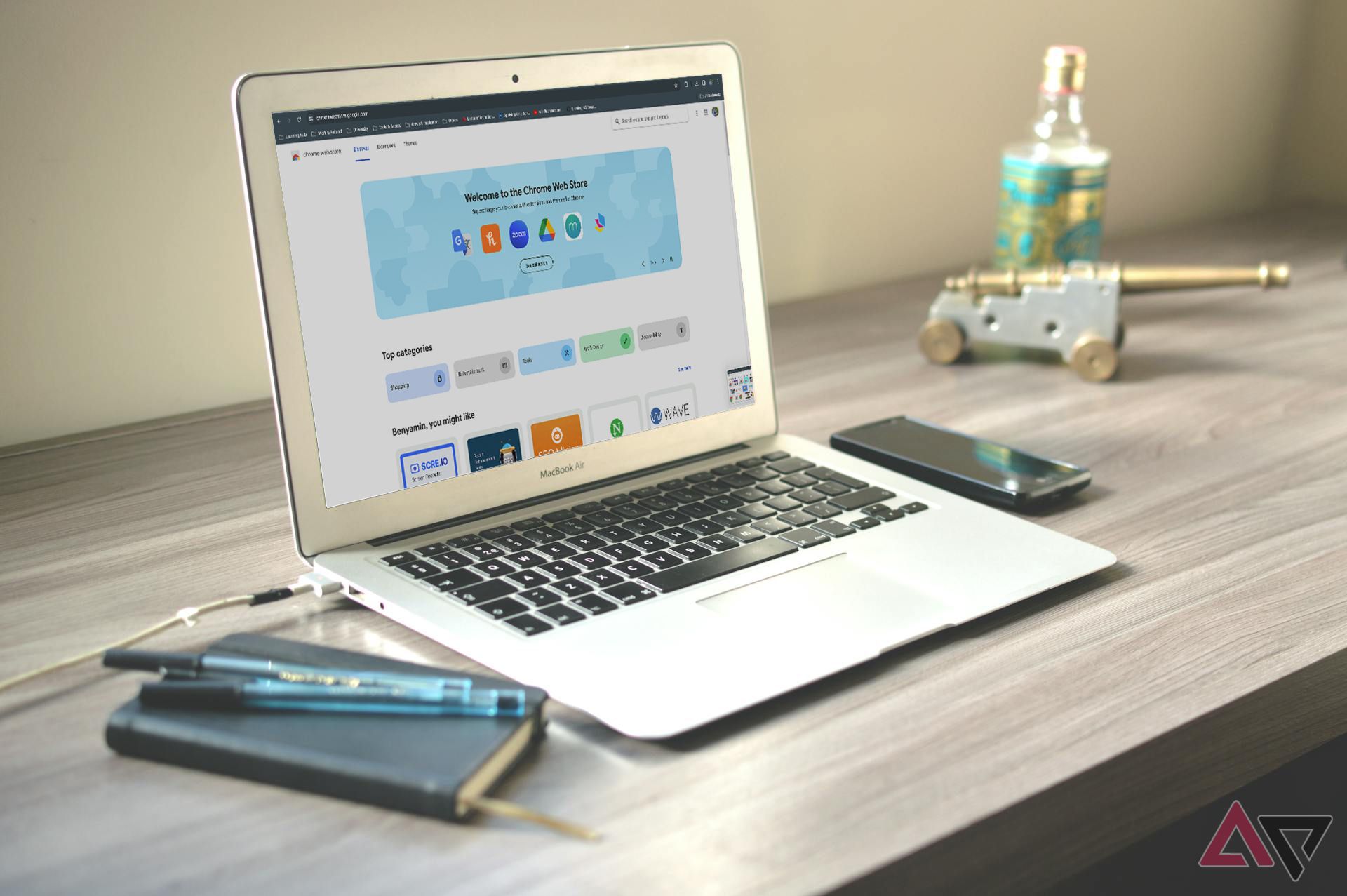 A laptop on a wooden desk displaying the Chrome Web Store homepage, with a smartphone, a bottle, pens, and a miniature cannon alongside.