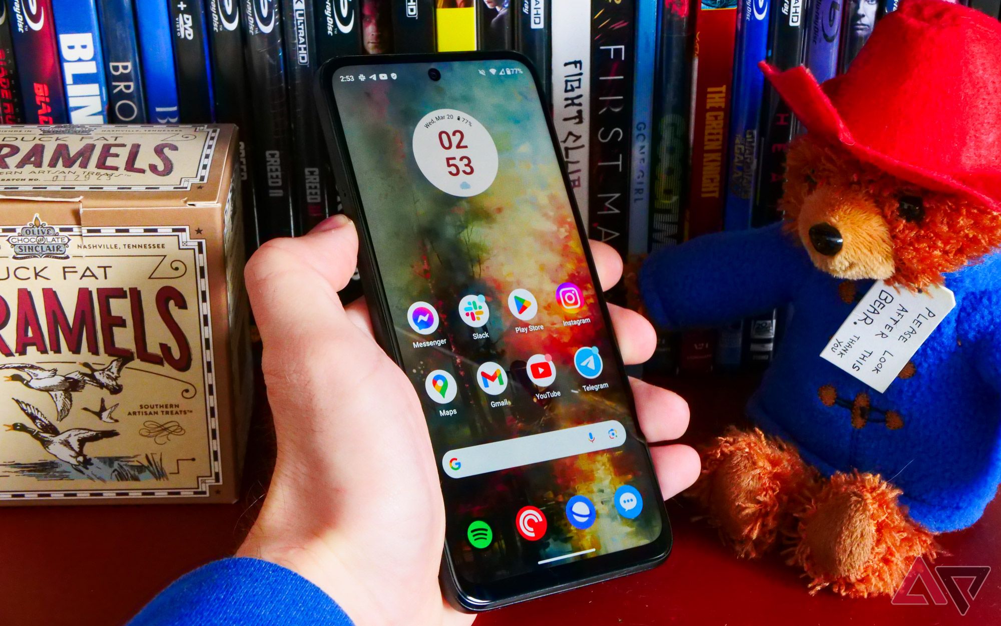 The Moto G Power (2024) held in a hand in front of a movie collection and next to a Paddington plushie.