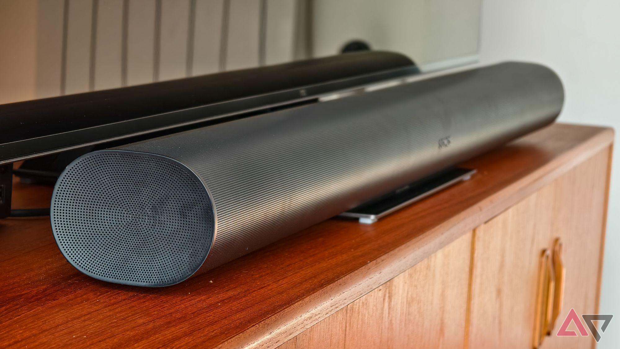 A black Sonos Arc soundbar positioned below a TCL TV on a teak wooden sideboard. The TV is off.