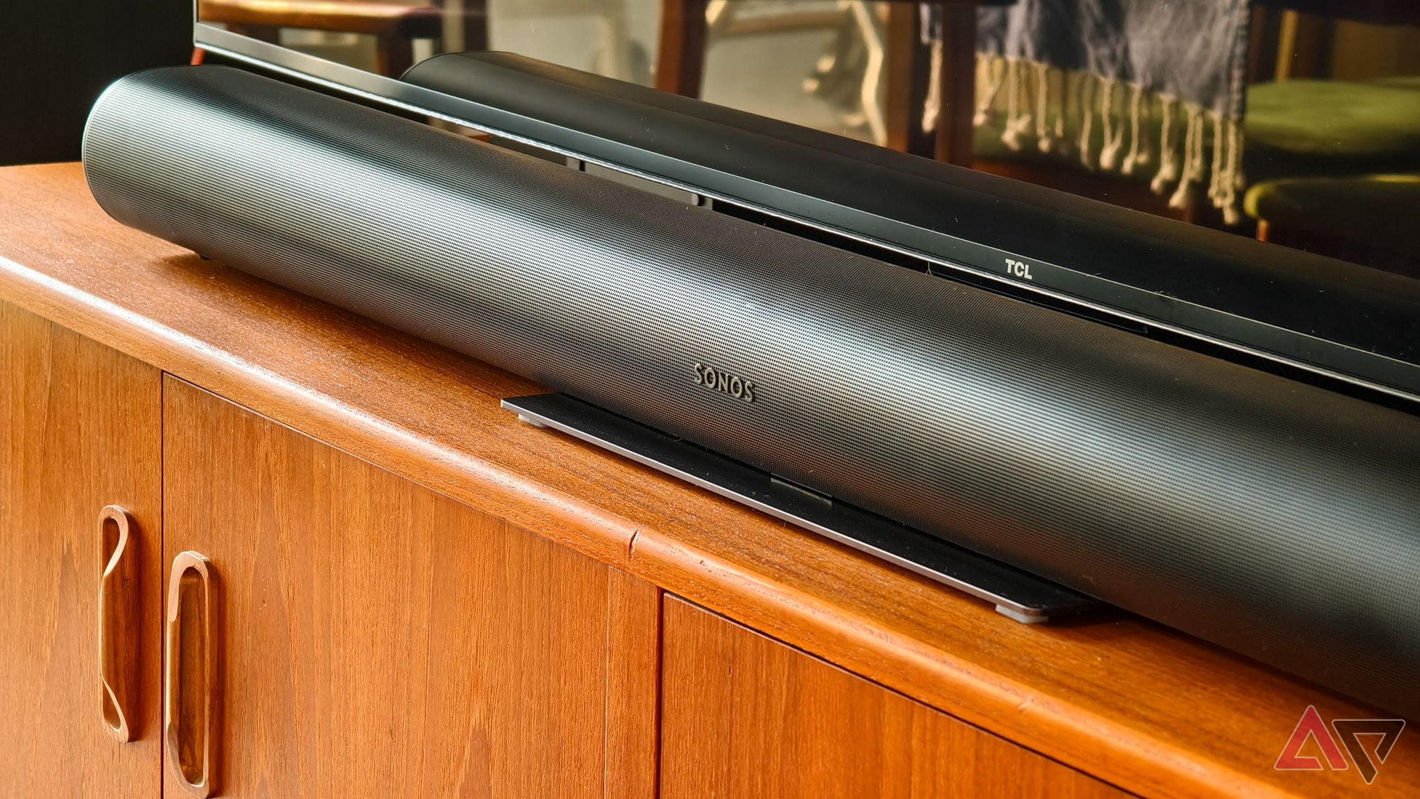 A photo of a black Sonos Arc soundbar on a teak wood TV stand, below a TCL TV, with the tassels of a blanket visible reflected in the TV