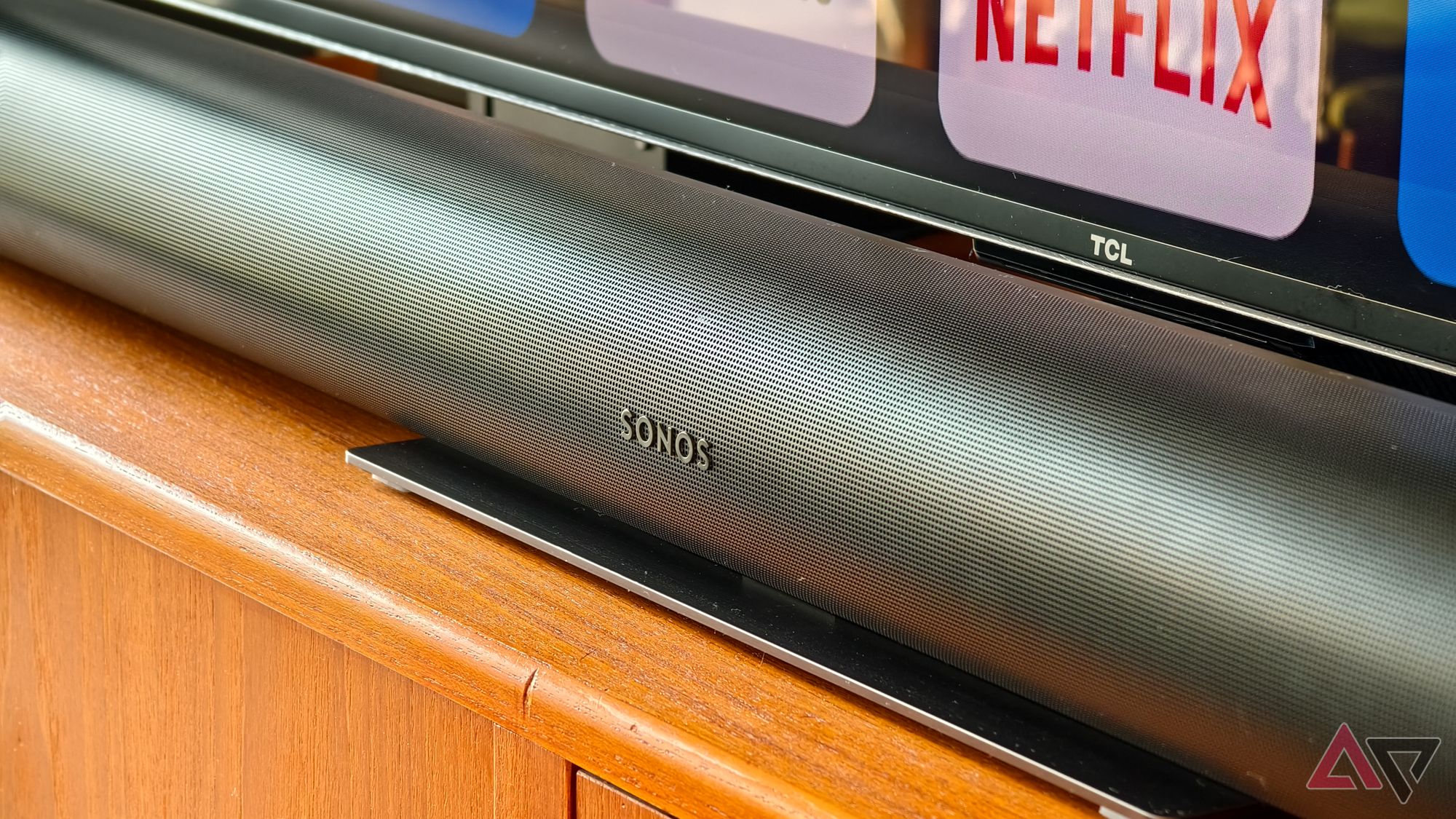 Sonos Arc soundbar in black, photographed above and from an angle, sitting on a teak TV stand with the bottom of the TCL TV visible behind showing a Netflix logo