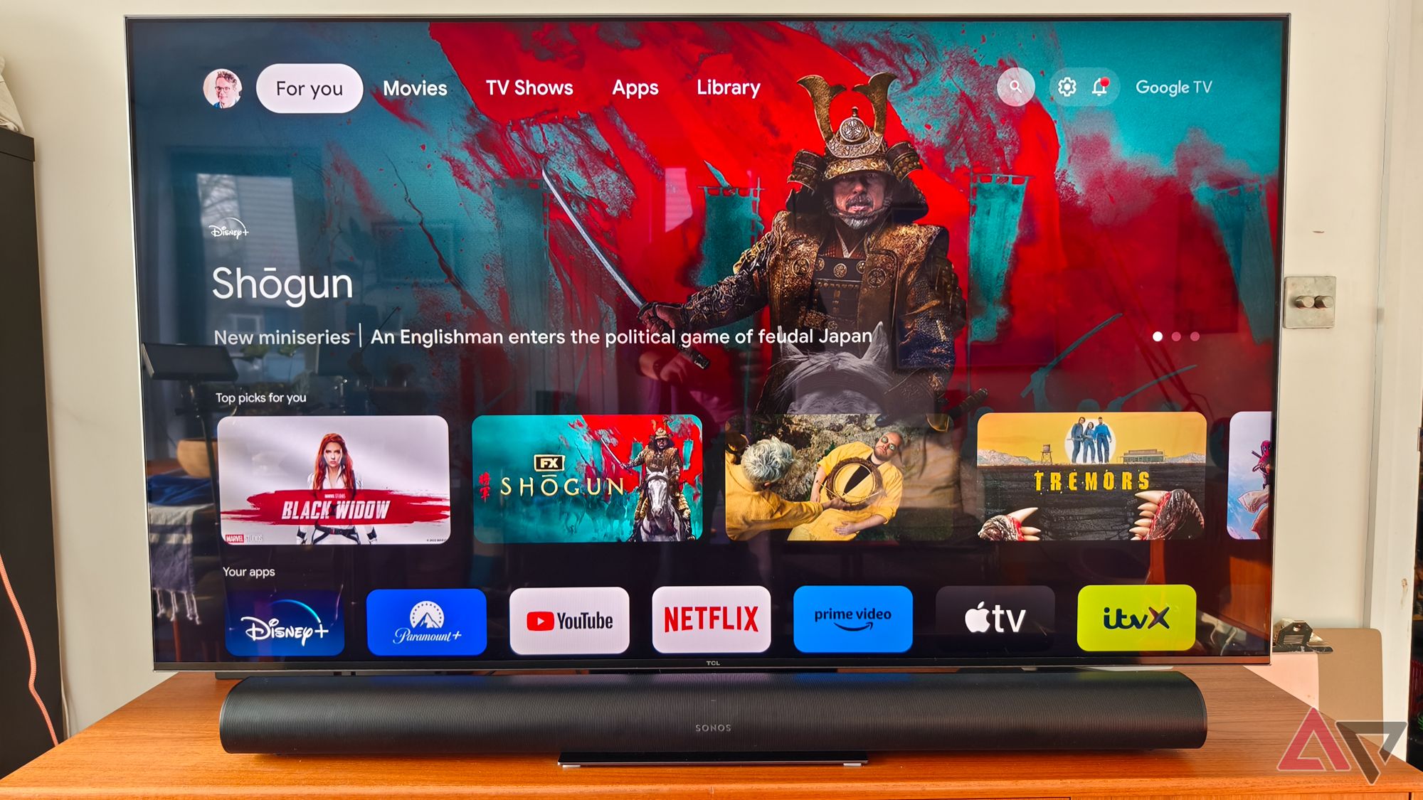 A head-on photo of a black Sonos Arc soundbar on a teak TV stand below a TCL TV. The TV is showing the Google TV home screen with a large ad for the TV series Shogun