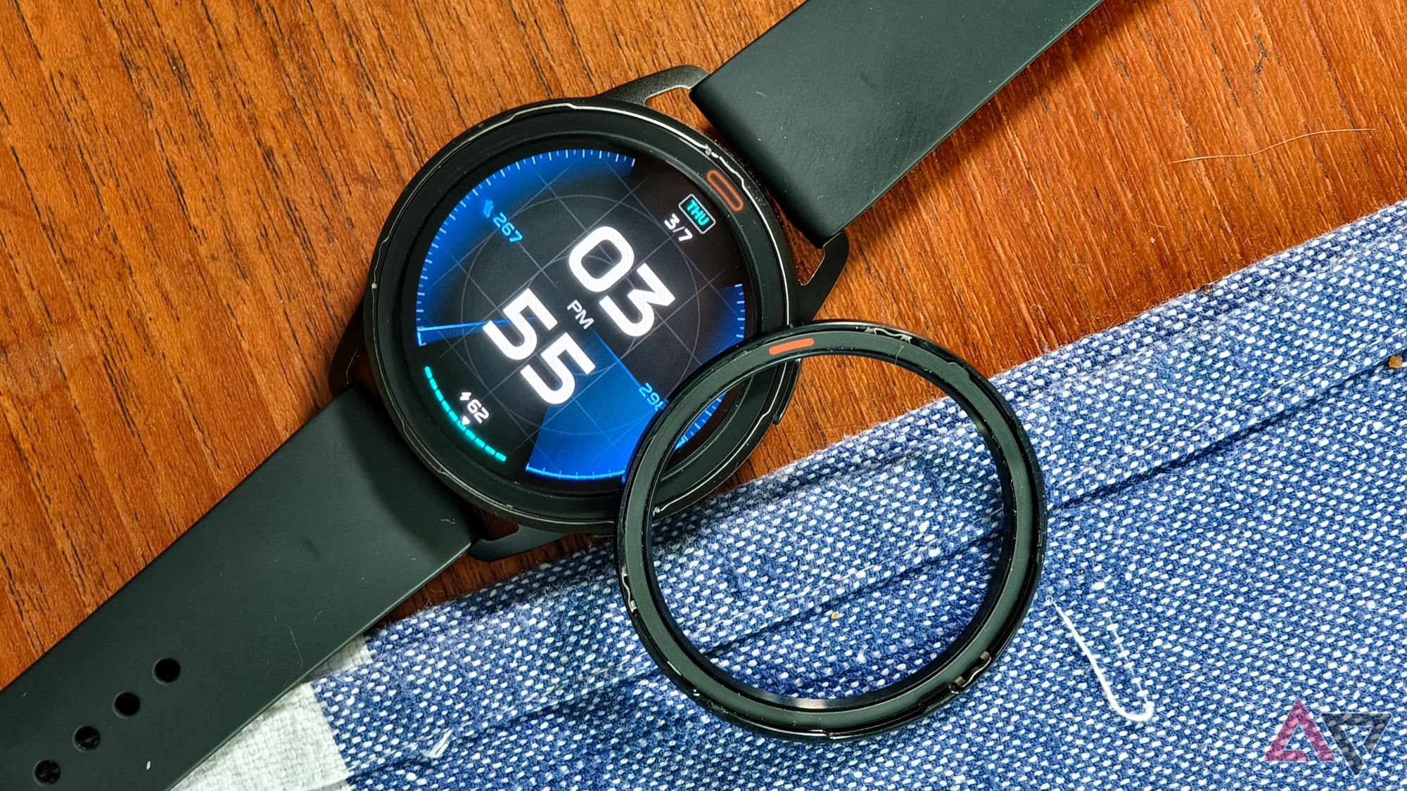 The Xiaomi Watch S3 viewed from above, lying on a teak wood table with blue fabric, with its bezel removed and lying upside down next to the watch