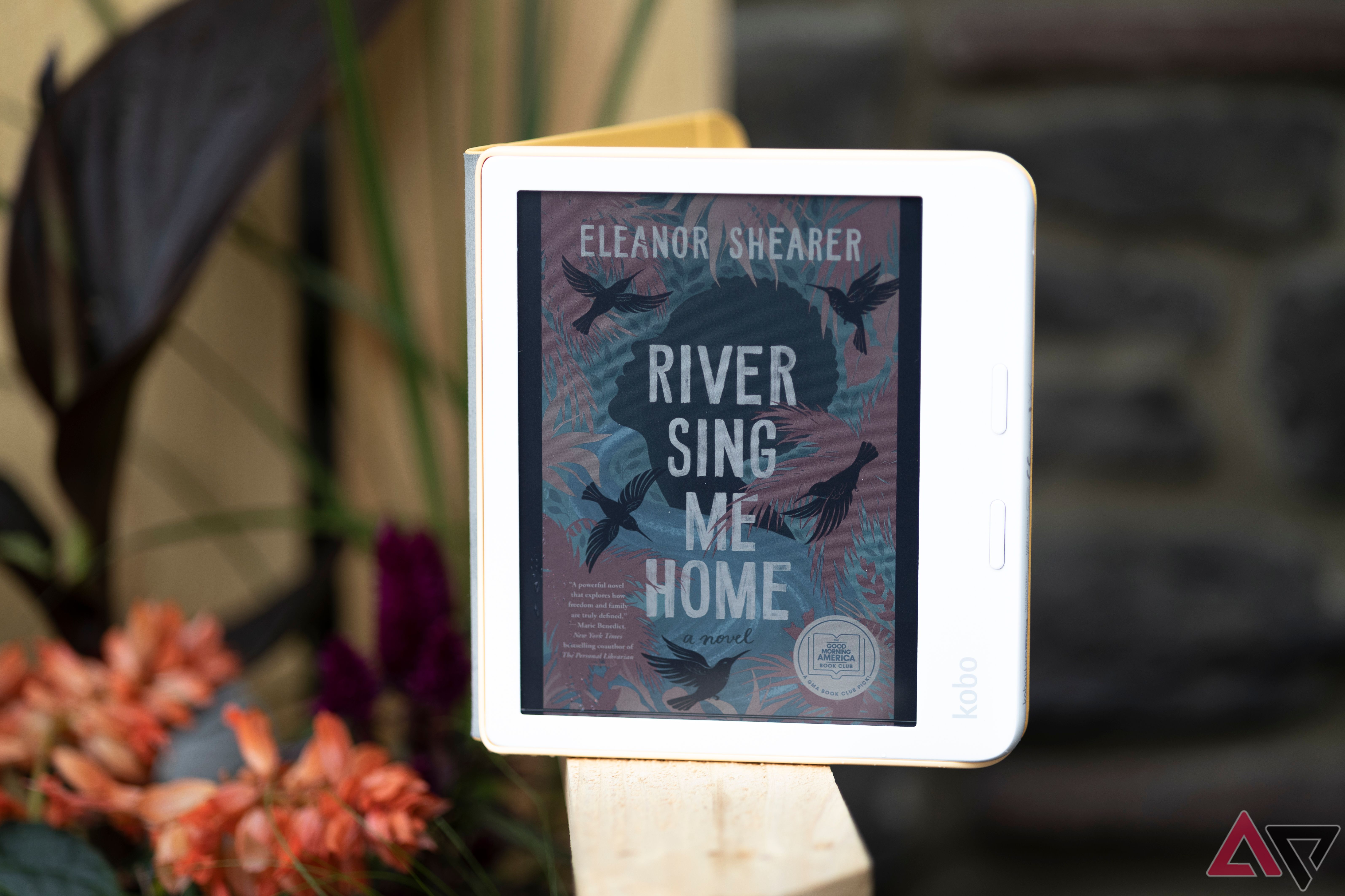 Kobo Libra Colour open to cover of 'River Sing Me Home' propped up on wooden bench