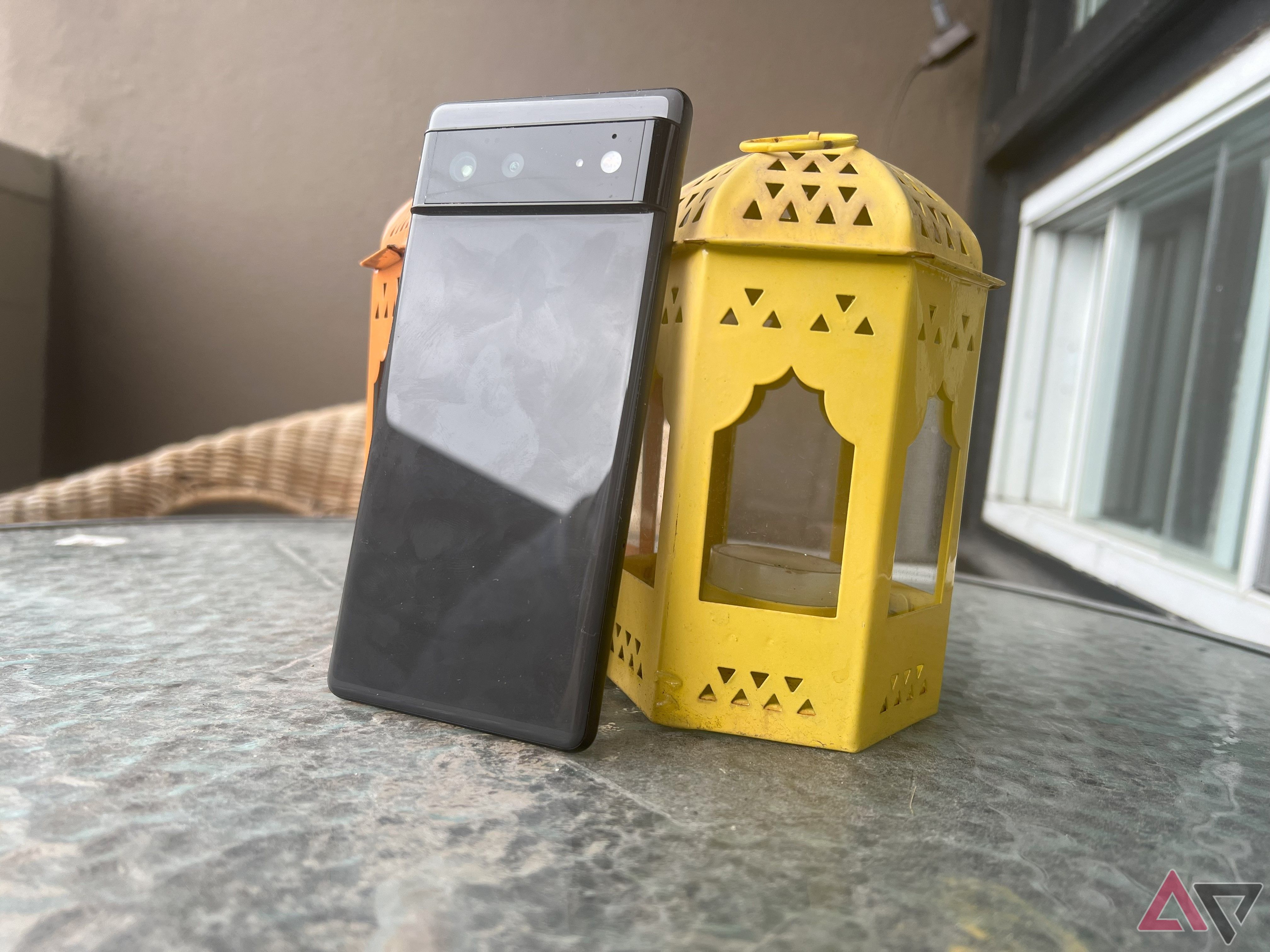 A Pixel 6 leaning on an orange and a yellow candle lantern on a patio table outdoors.