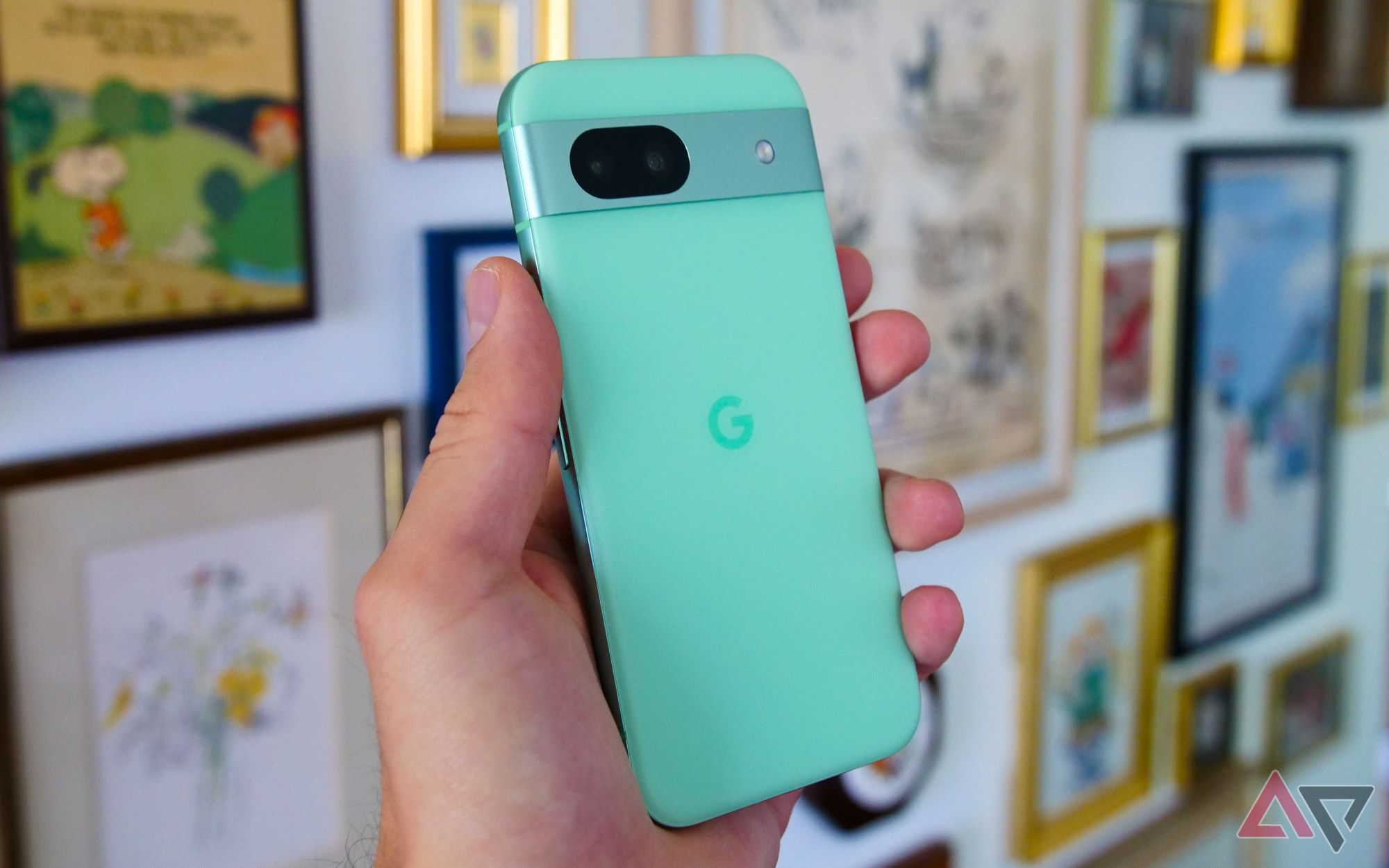 The Google Pixel 8a held in a hand with photos hung on the wall behind it.