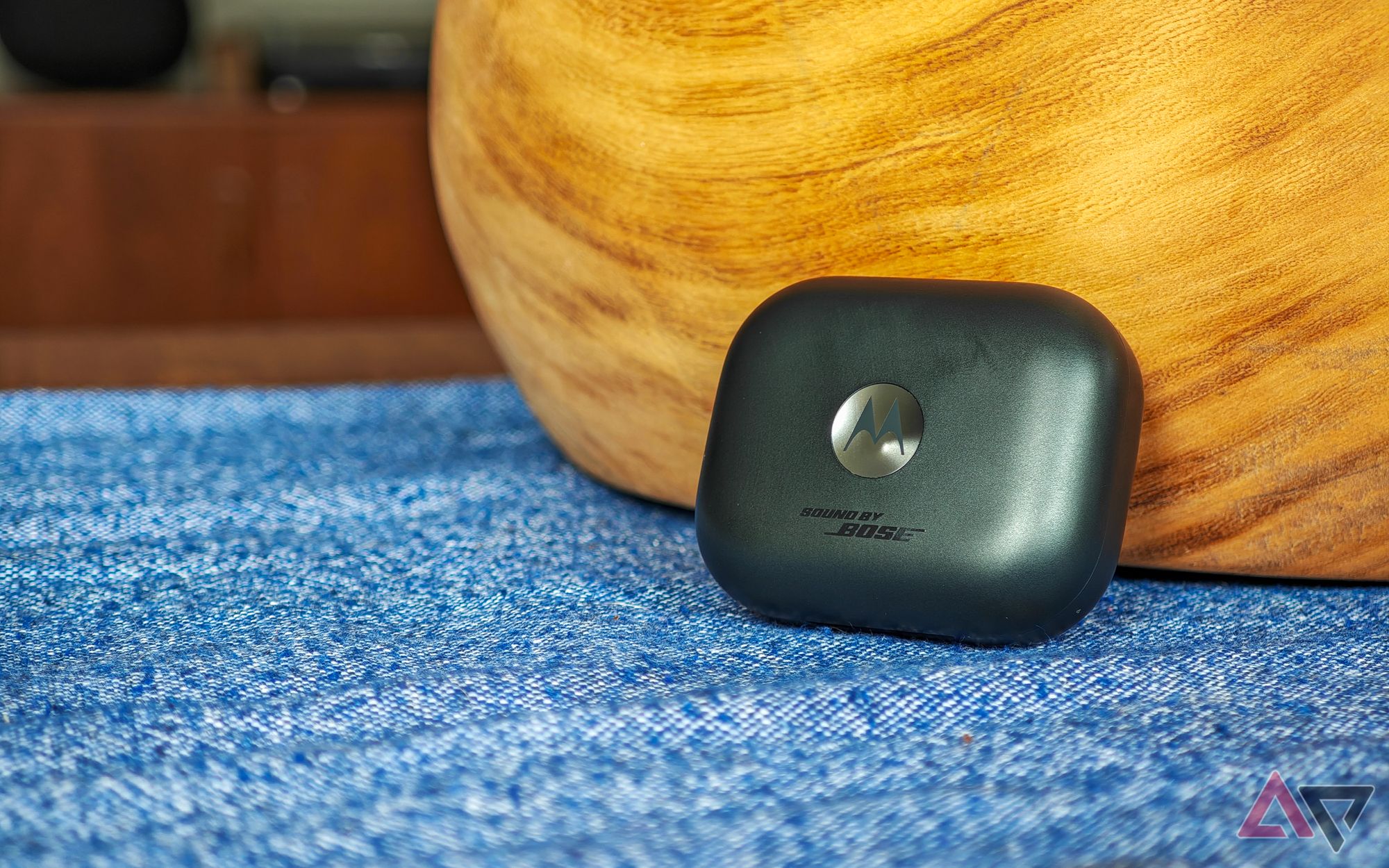 The Moto Buds+ in black resting against a wooden bowl on a blue tablecloth