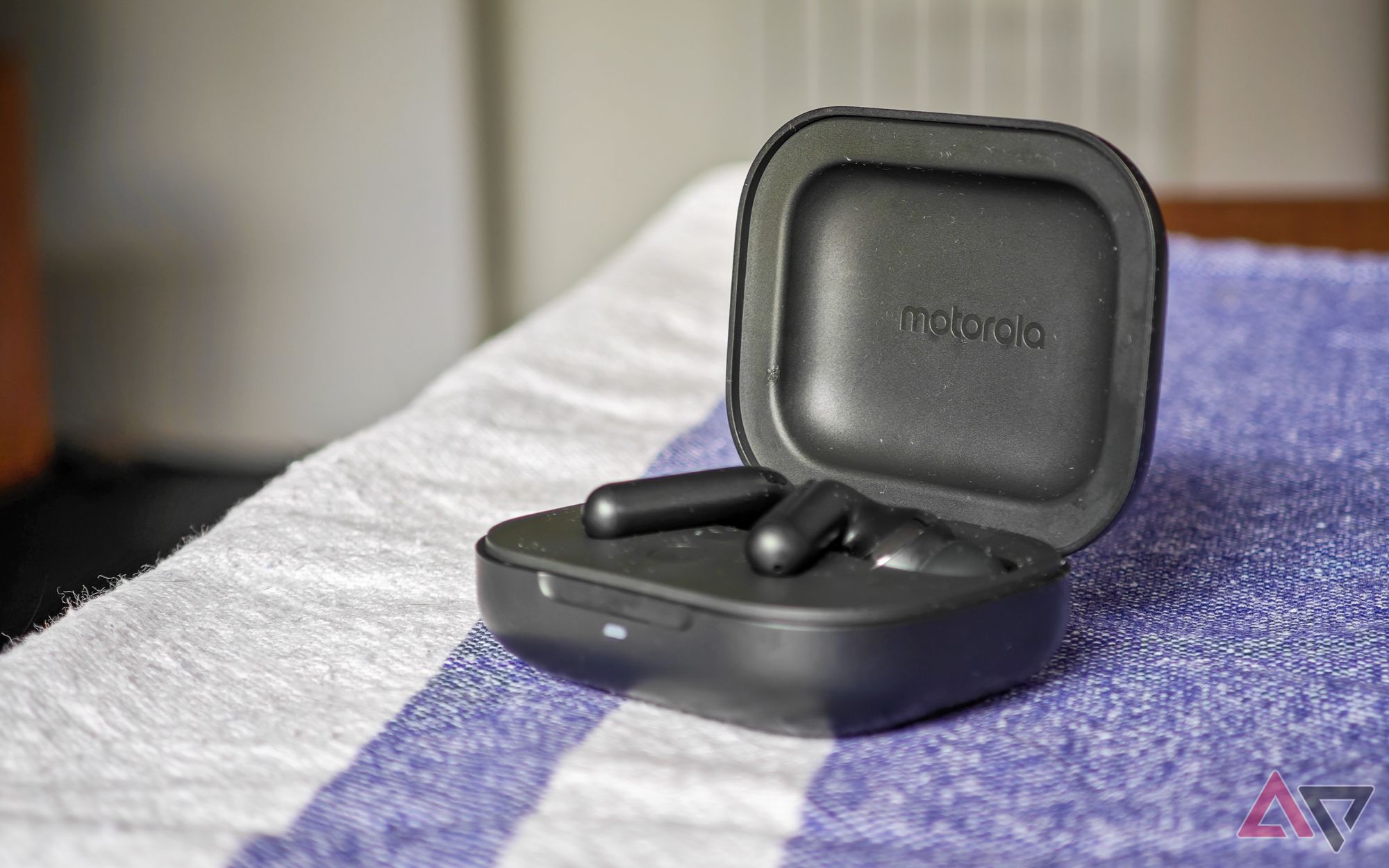 The Moto Buds+ in black with the earbuds in the open charging case on a blue and white tablecloth