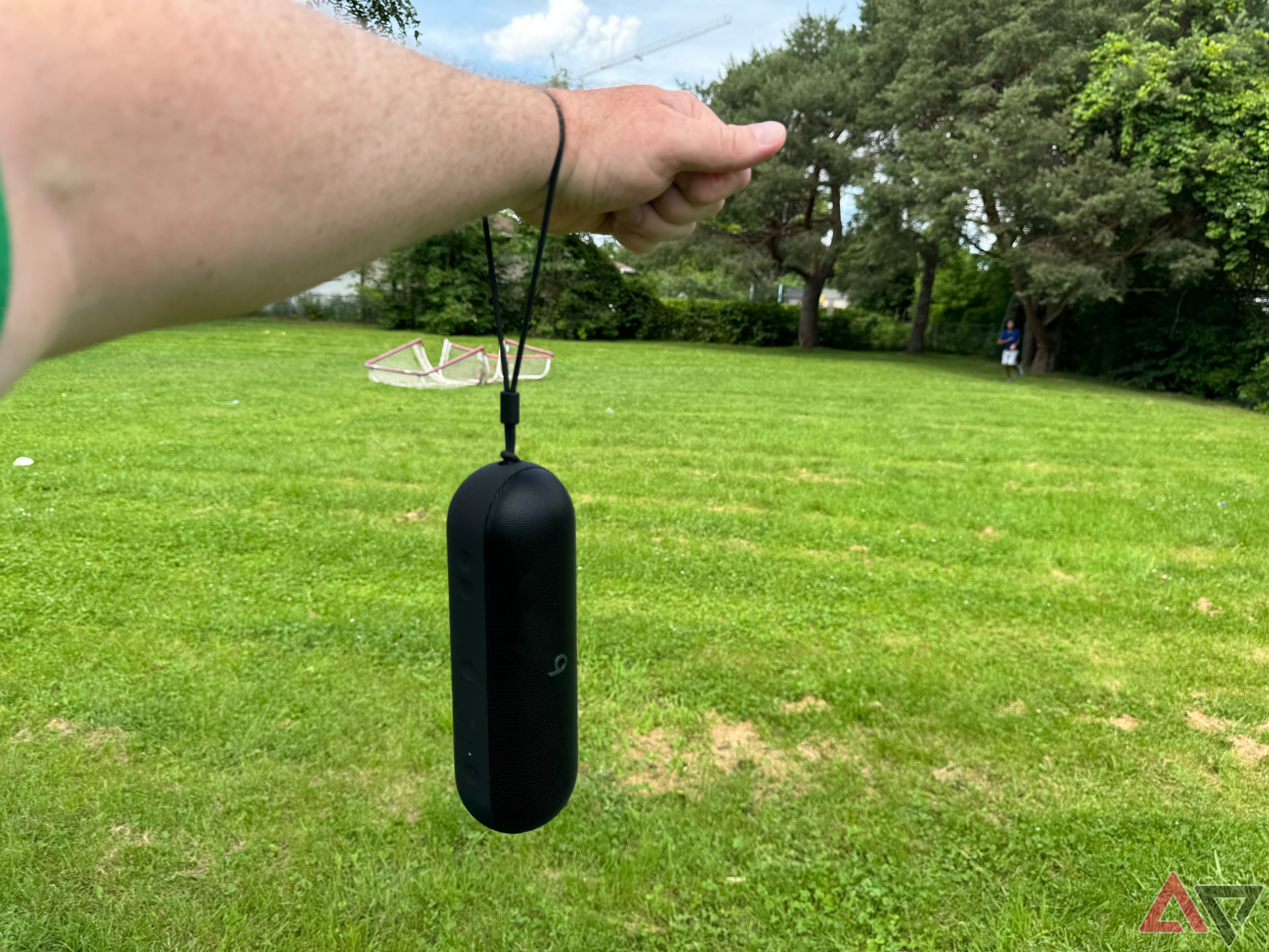 A black 2024 Beats Pill dangling on its lanyard from an oustretched arm above a green grassy field.