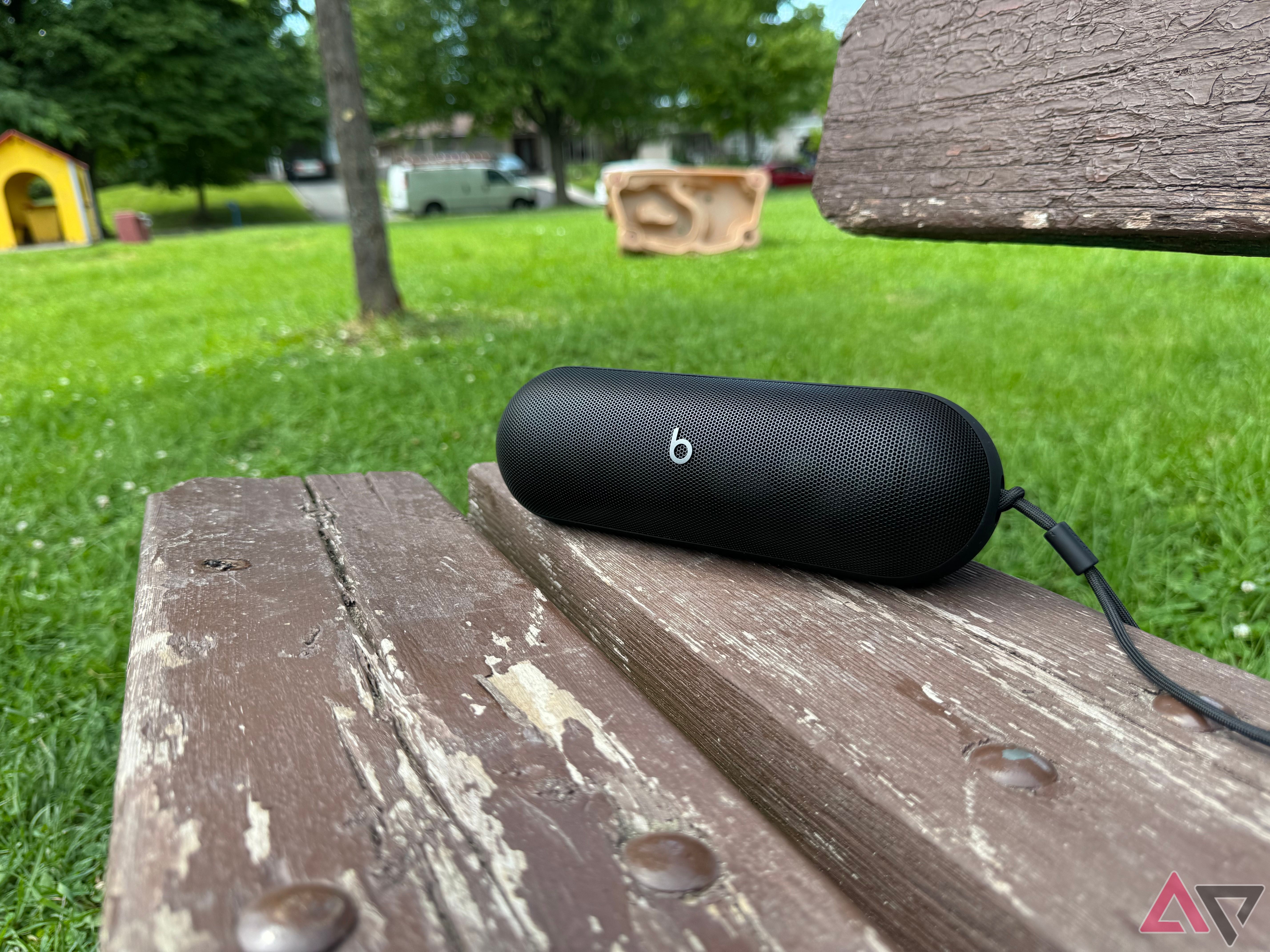 A black 2024 Beats Pill on a brown wooden park bench with green grass and trees in the background.