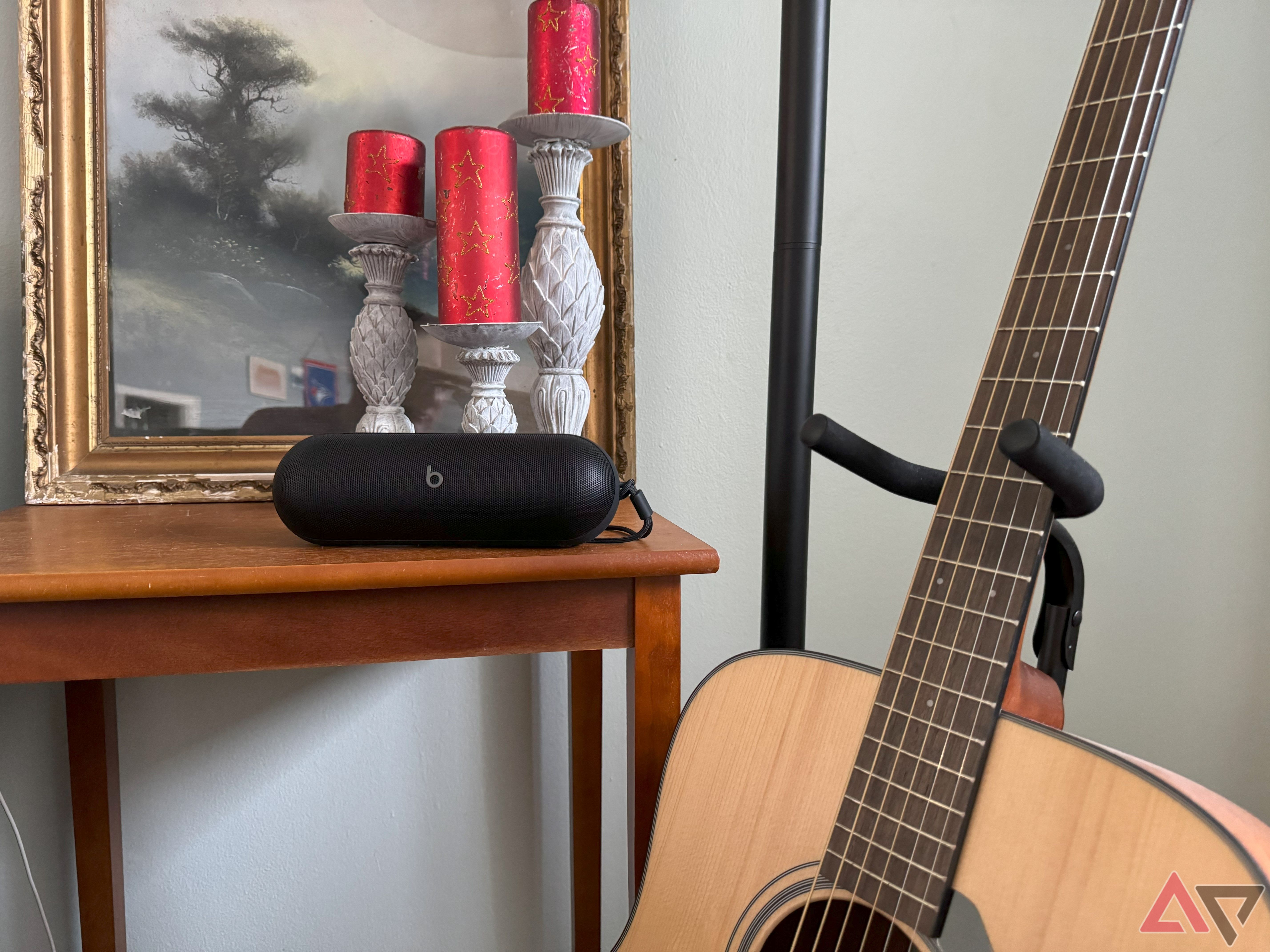 A black 2024 Beats Pill on a wooden side table with three red candles behind it next to an acoustic guitar. 