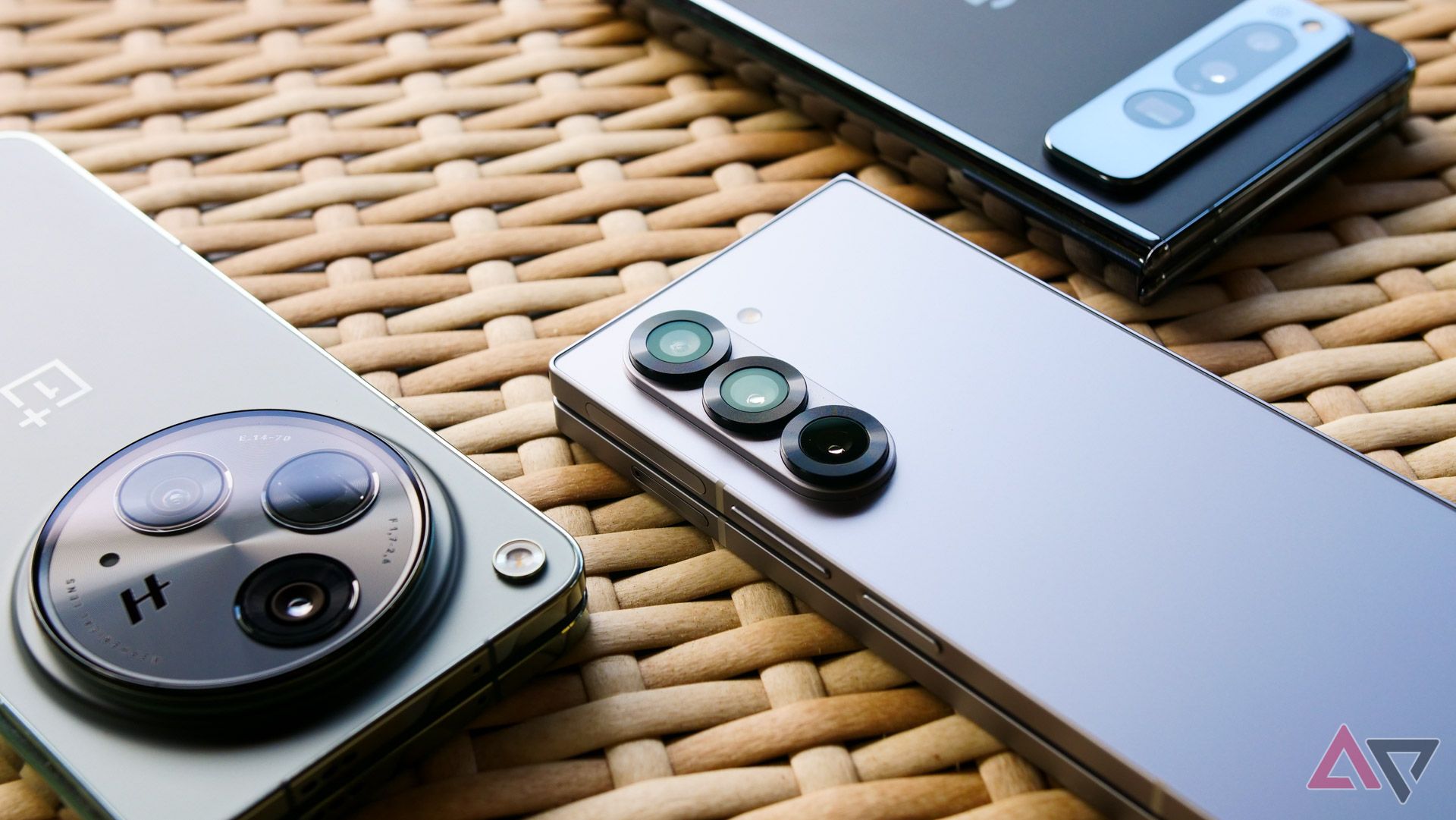 The Galaxy Z Fold 6, OnePlus Open, and Pixel Fold sitting on a wicker table with their camera bumps shown.