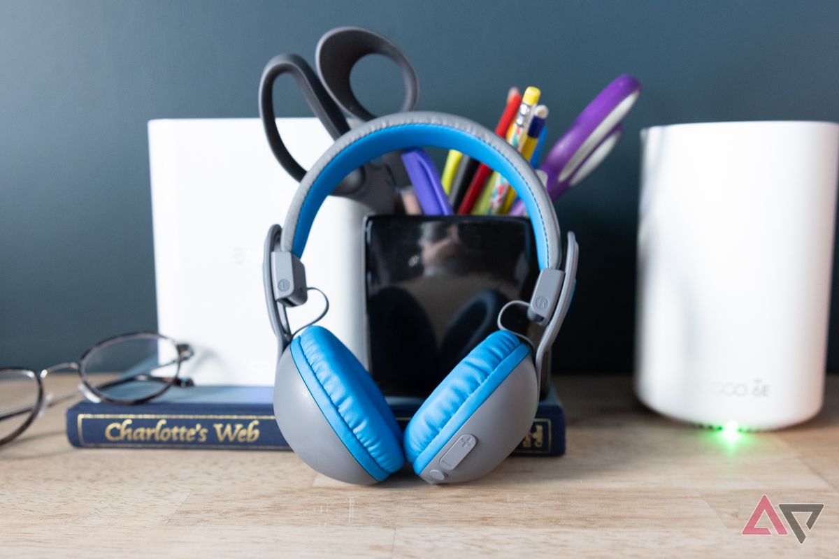 JLab JBuddies Studio 2 headphones leaning against a book, pens, routers, and near some eyeglasses