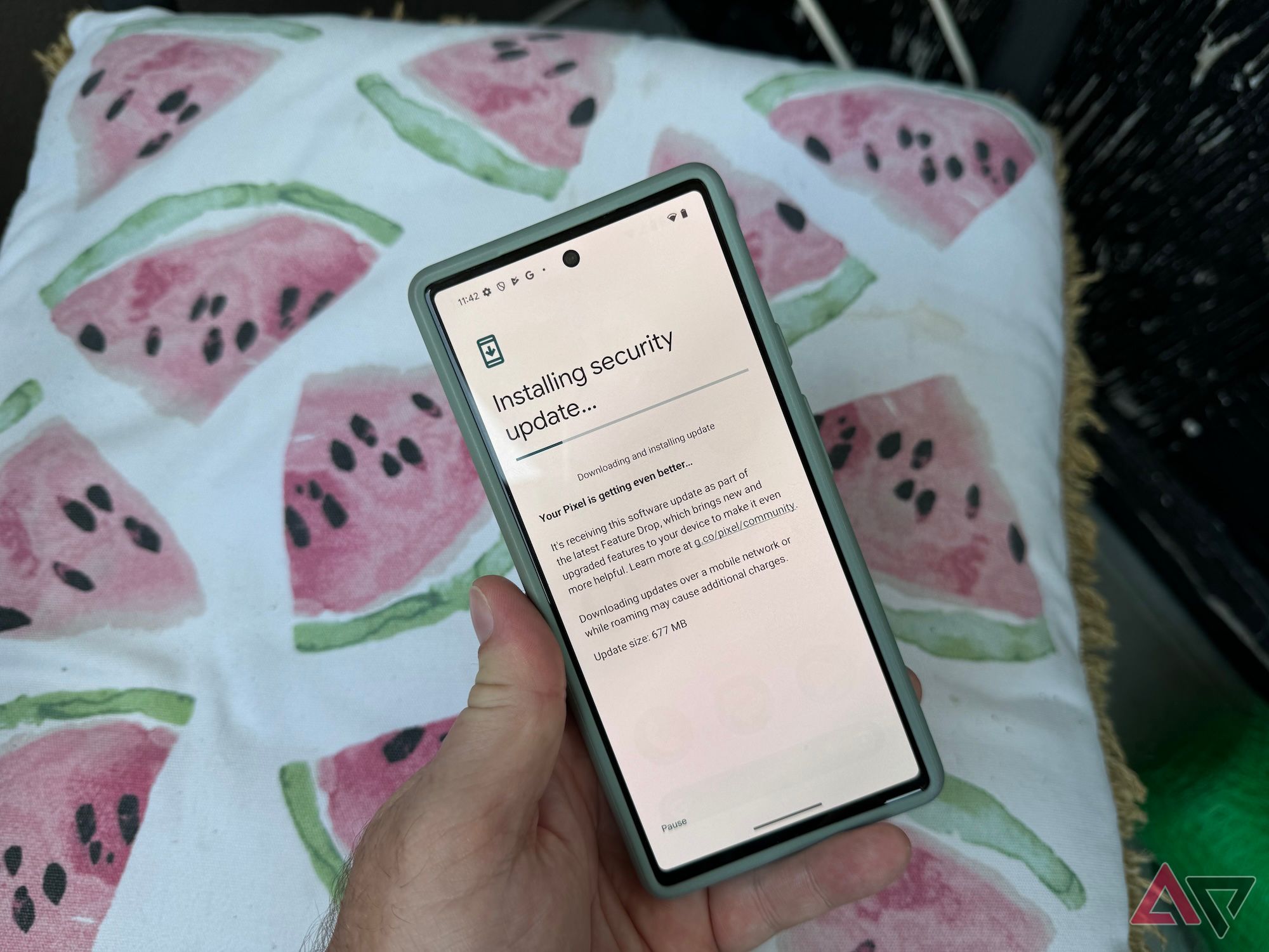 A man's hand holding a Pixel 6 showing an update in progress with a pillow in the background that has pictures of pink watermelon slices on it.