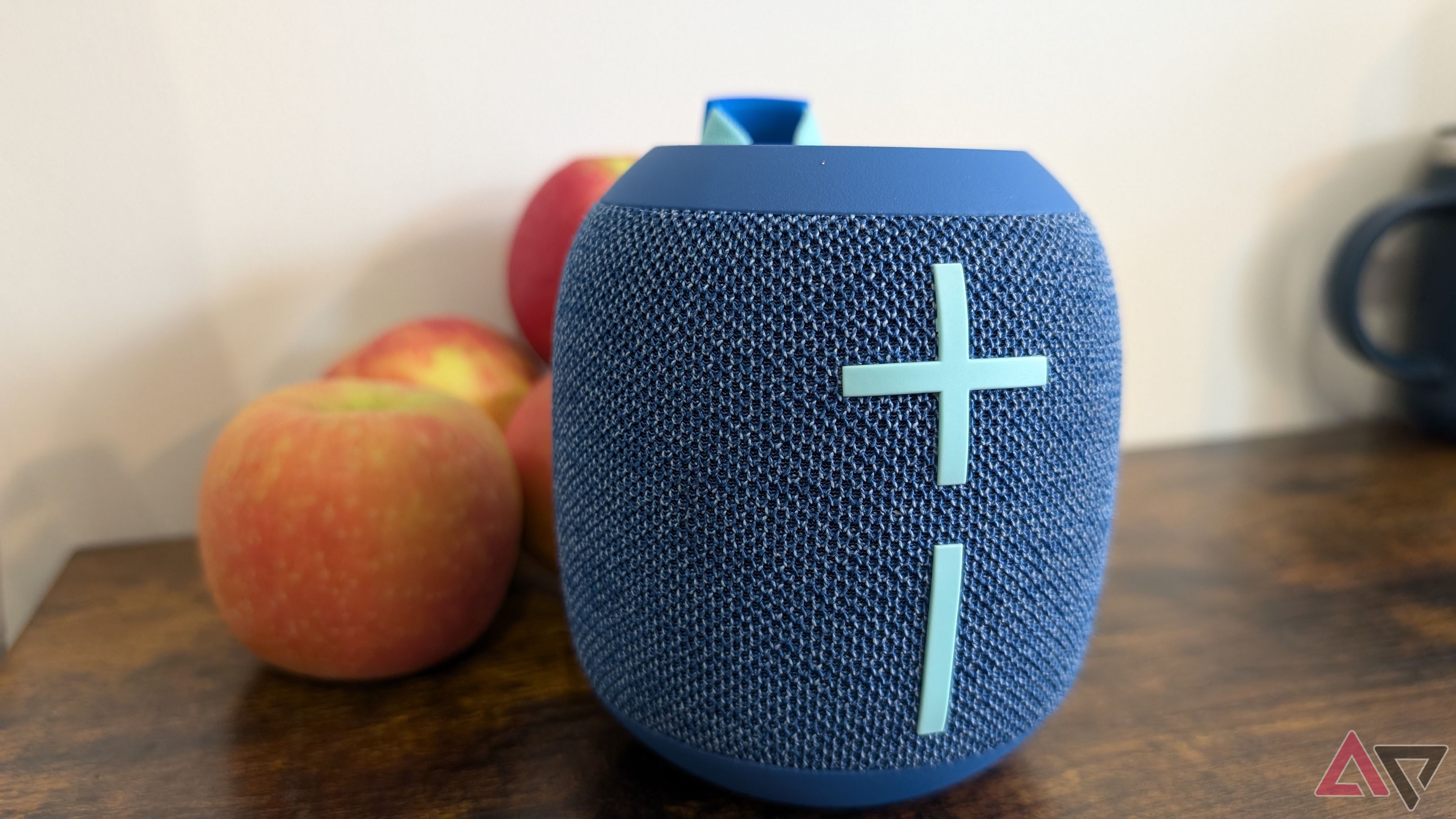 blue bluetooth speaker with light blue plus and minus buttons on wood table in front of apples