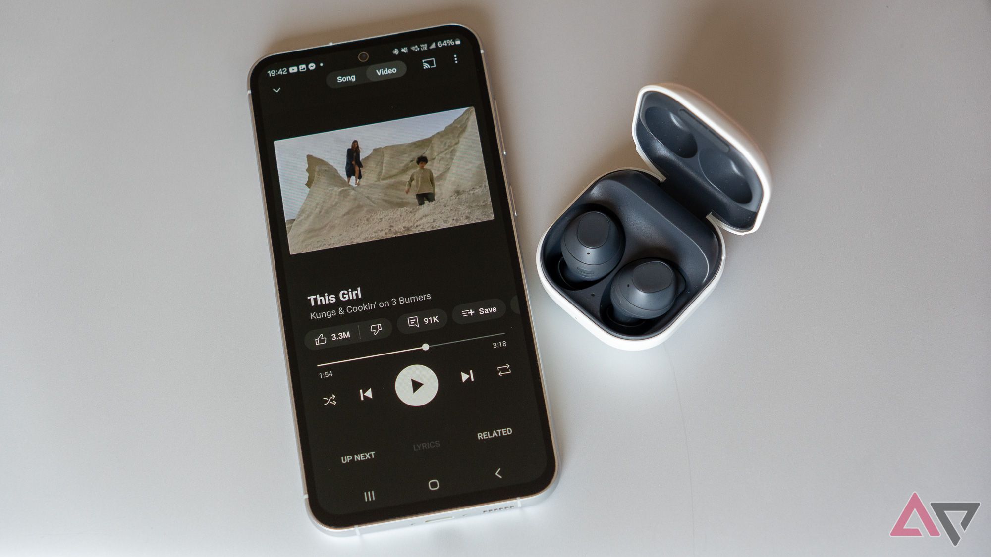 YouTube Music Premium Playing On A Samsung Phone Next To Samsung Wireless Earbuds On A Gray Metal Surface.