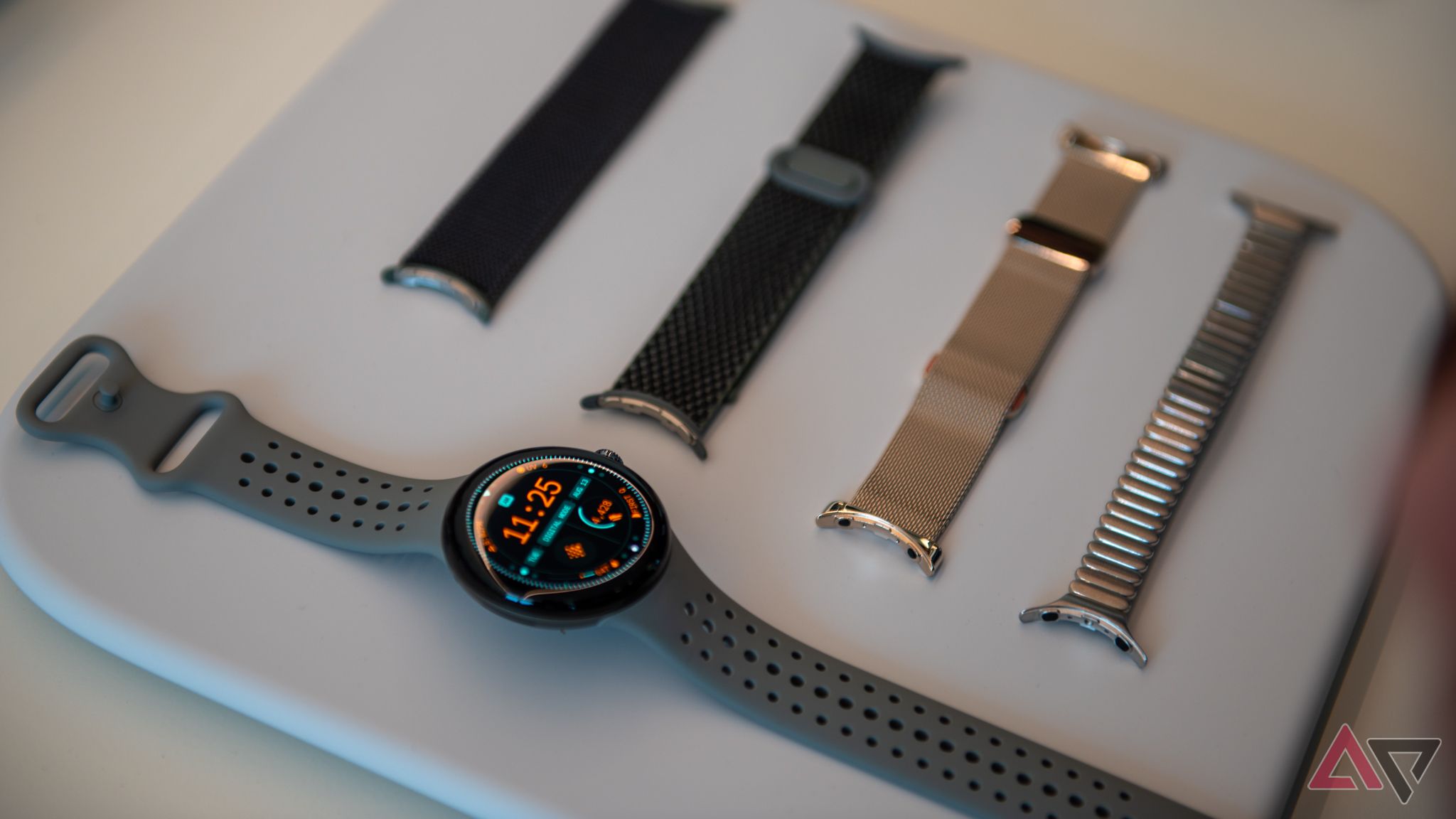 A Google Pixel Watch next to a selection of four watch bands