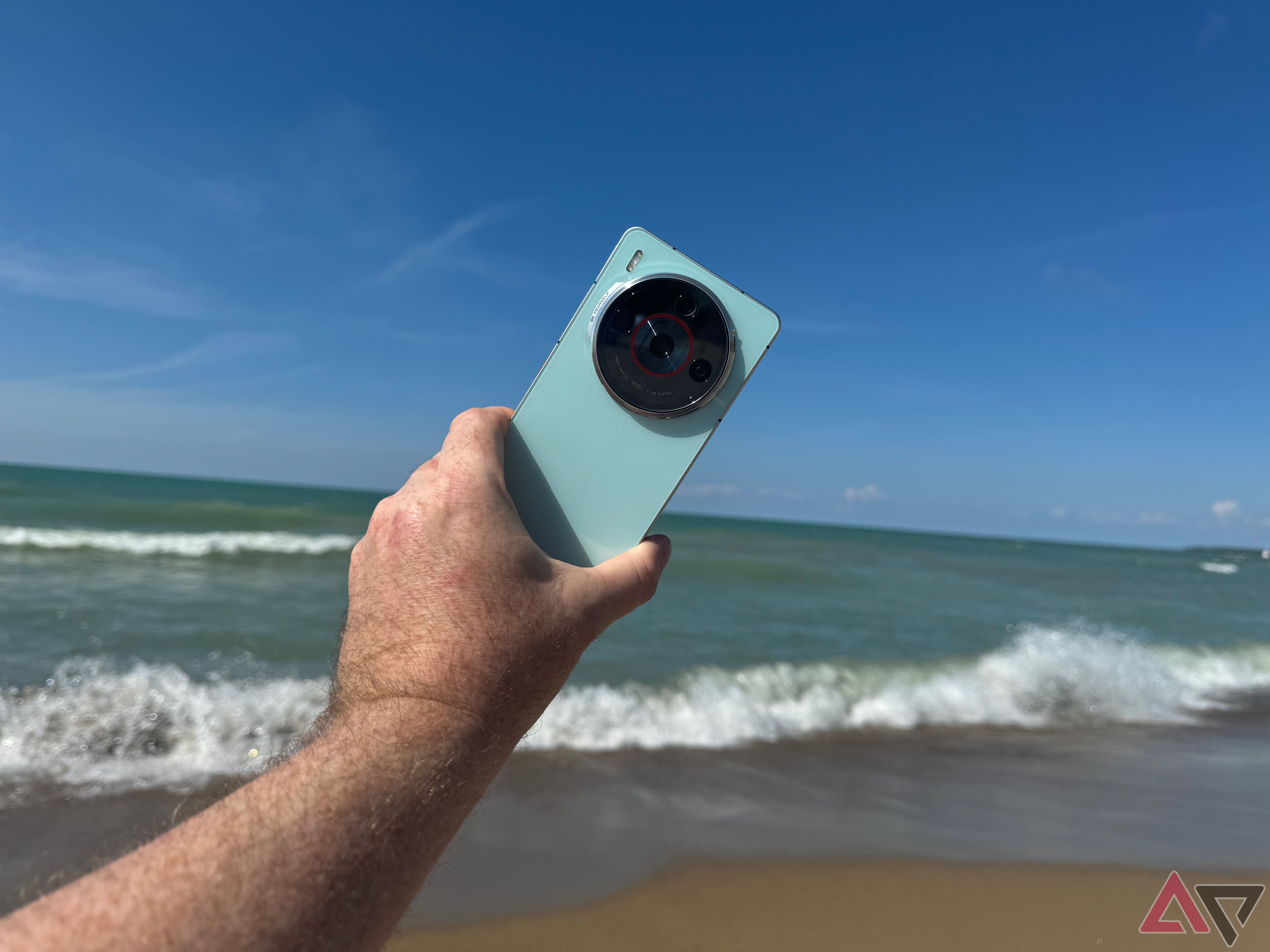 A man's hand holding an aqua Nubia Z60S Pro in the air on a beach with waves in the background.