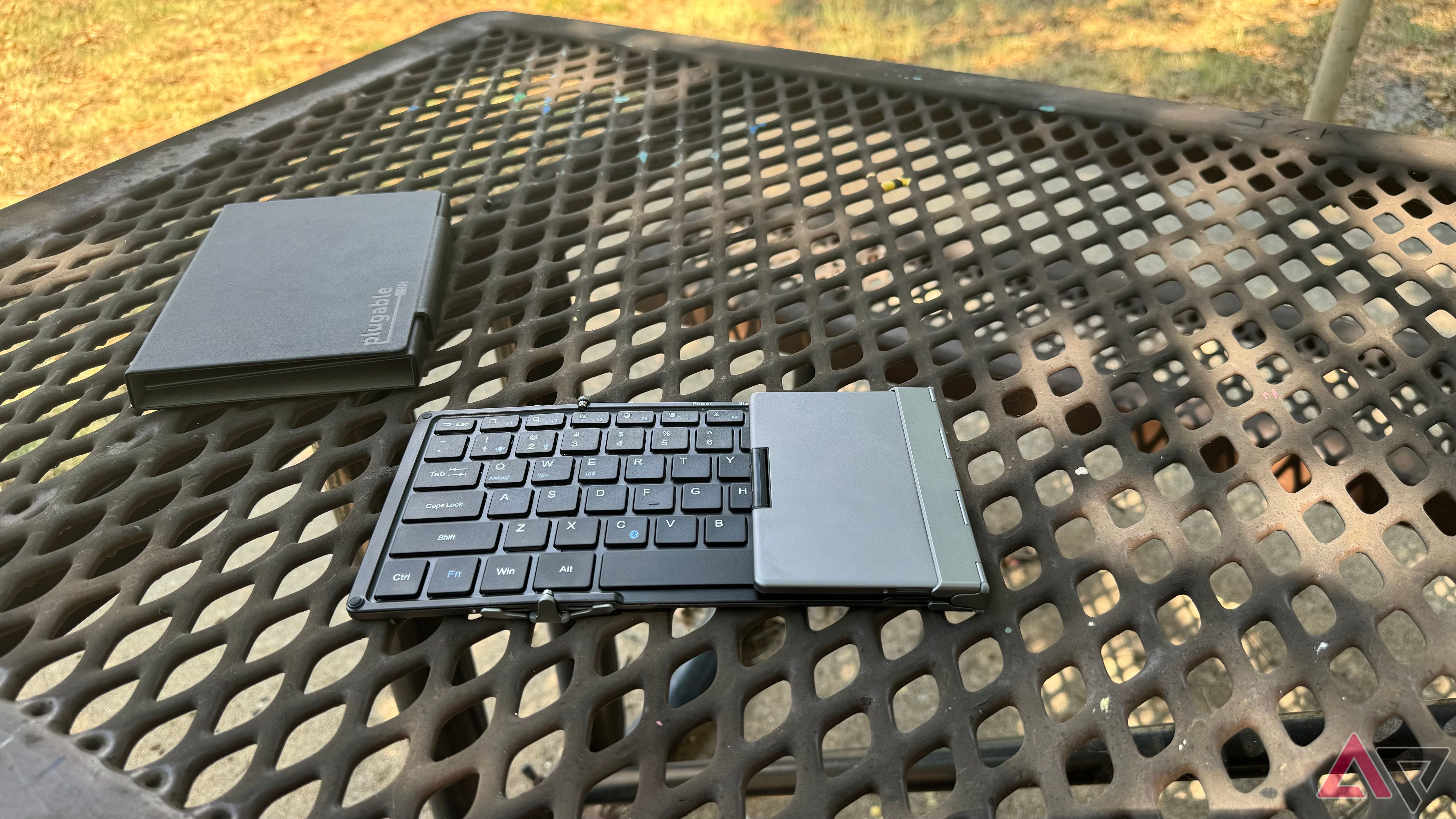 Plugable Foldable Bluetooth Keyboard half-folded sitting next to carrying case on a shaded picnic table