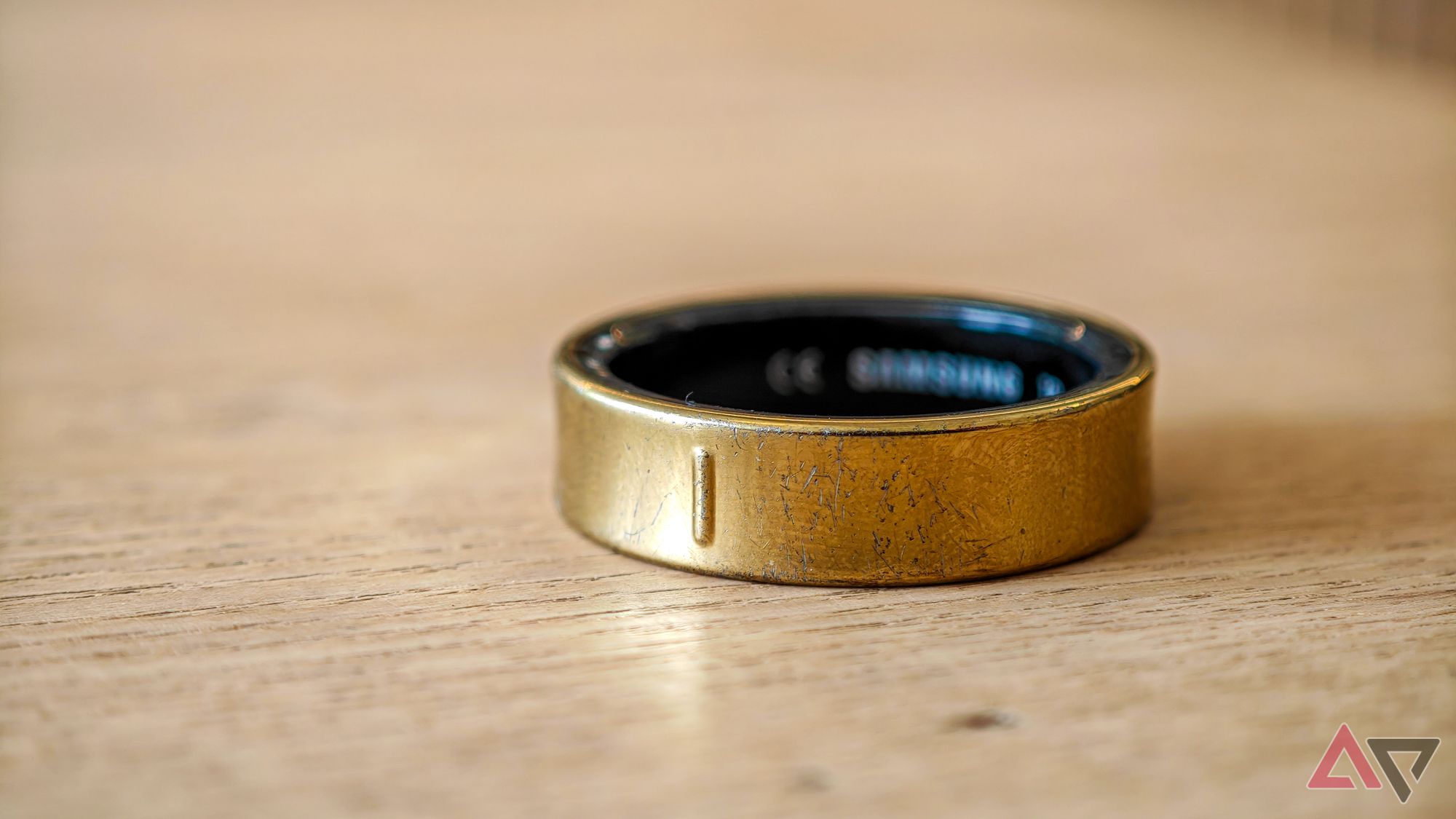 A gold Samsung Galaxy Ring lying on its side on a pale wooden table with some visible scratches to the finish