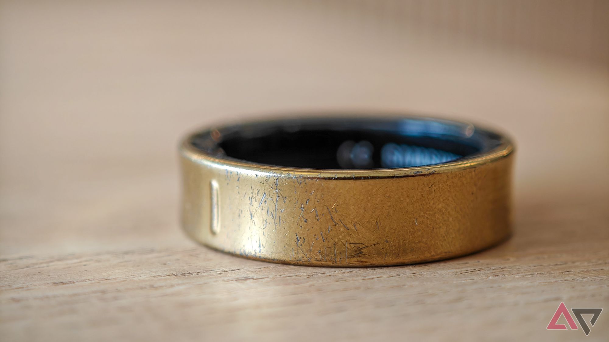 A gold Samsung Galaxy Ring on its side on a pale wooden table with a close-up on its clear scratches