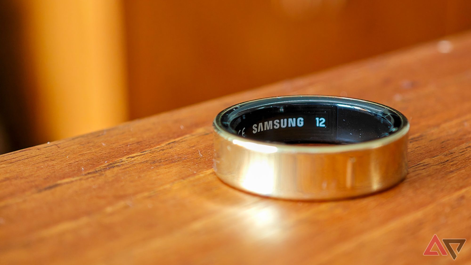 A gold Samsung Galaxy Ring resting on its side on a wooden surface, with the focus on the Samsung logo on the ring's inside