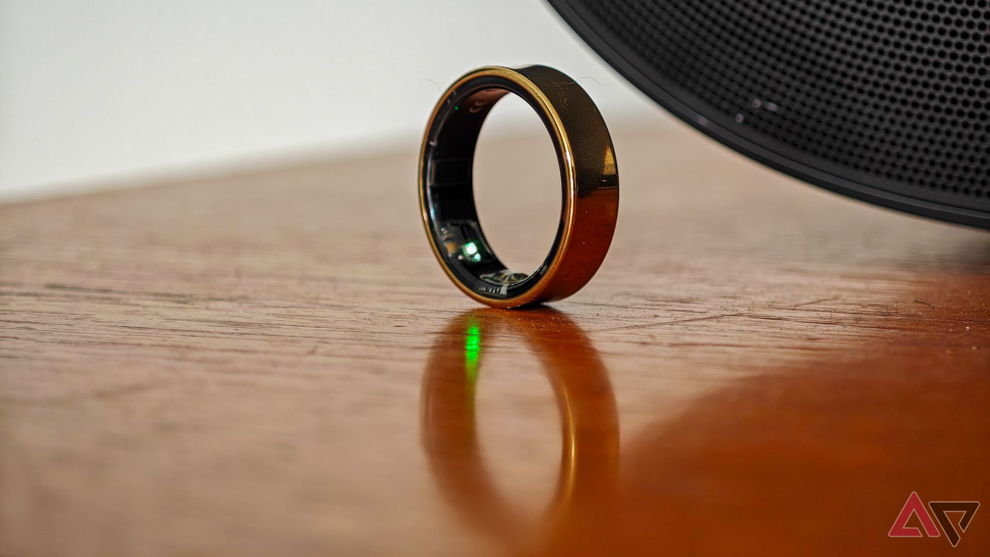 A gold Samsung Galaxy Ring standing on its side on a wooden surface, with a green tracker LED lit