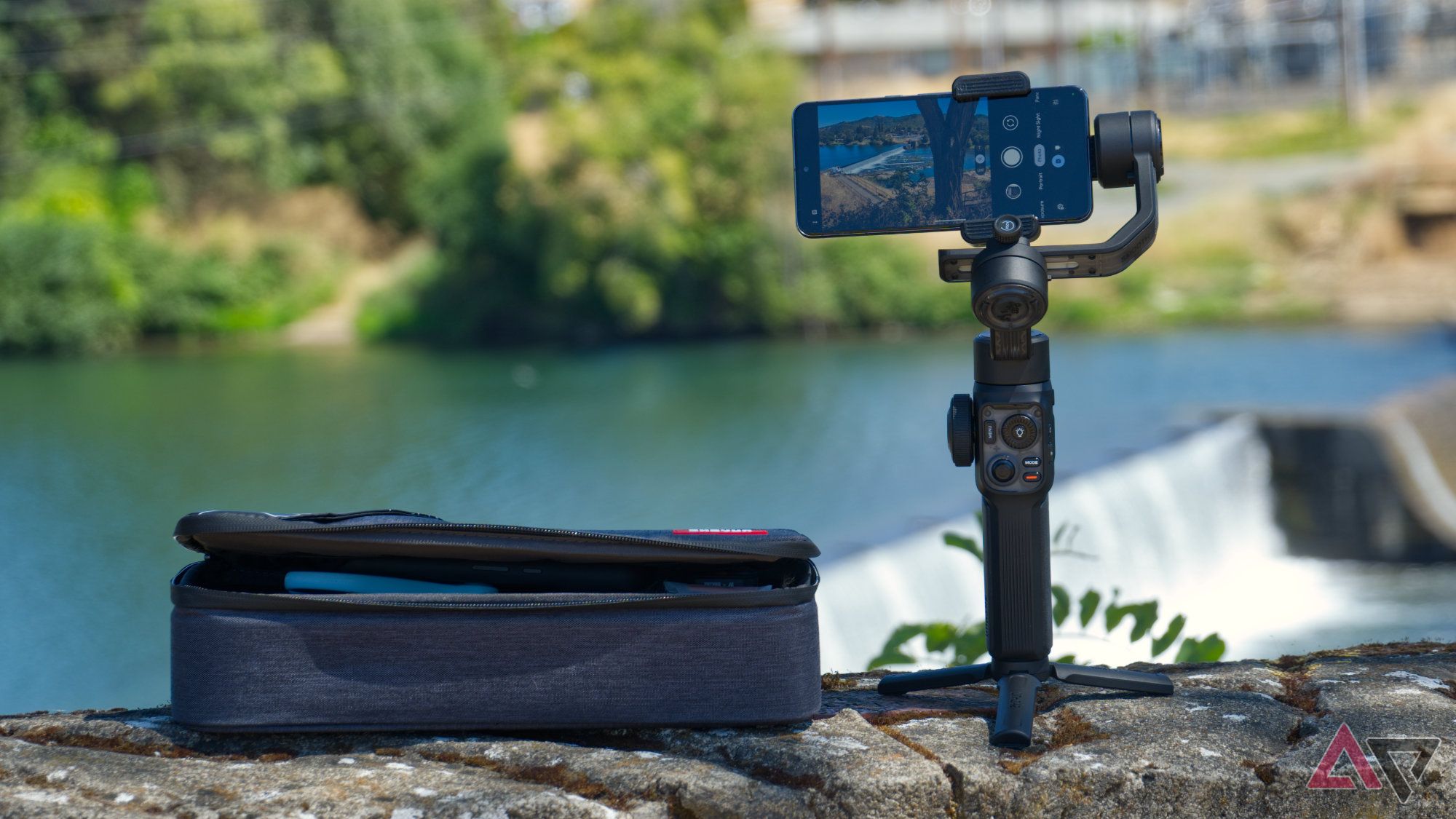 Zhiyun Smooth 5S AI gimbal standing upright on its tripod next to the travel case, overlooking a dam