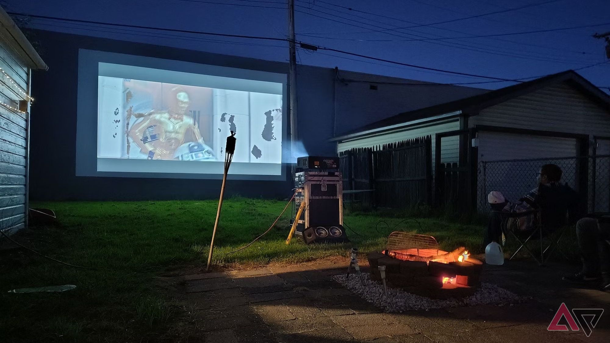 A scene taken outdoors showcasing a fire and a movie playing on a projector.