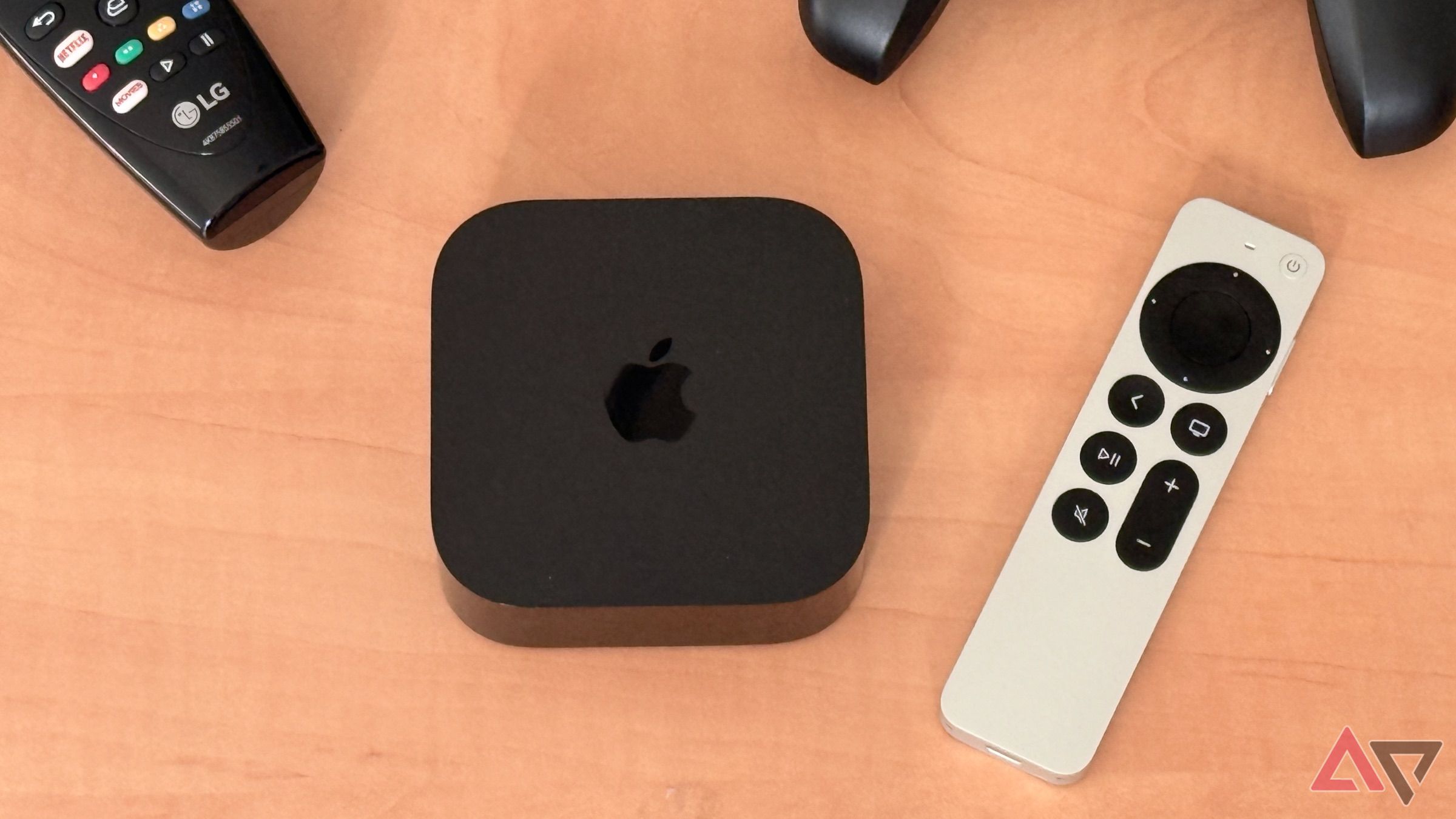 Apple TV 4K on light wooden desk with Siri Remote, LG TV remote, and Nimbus Game Controller