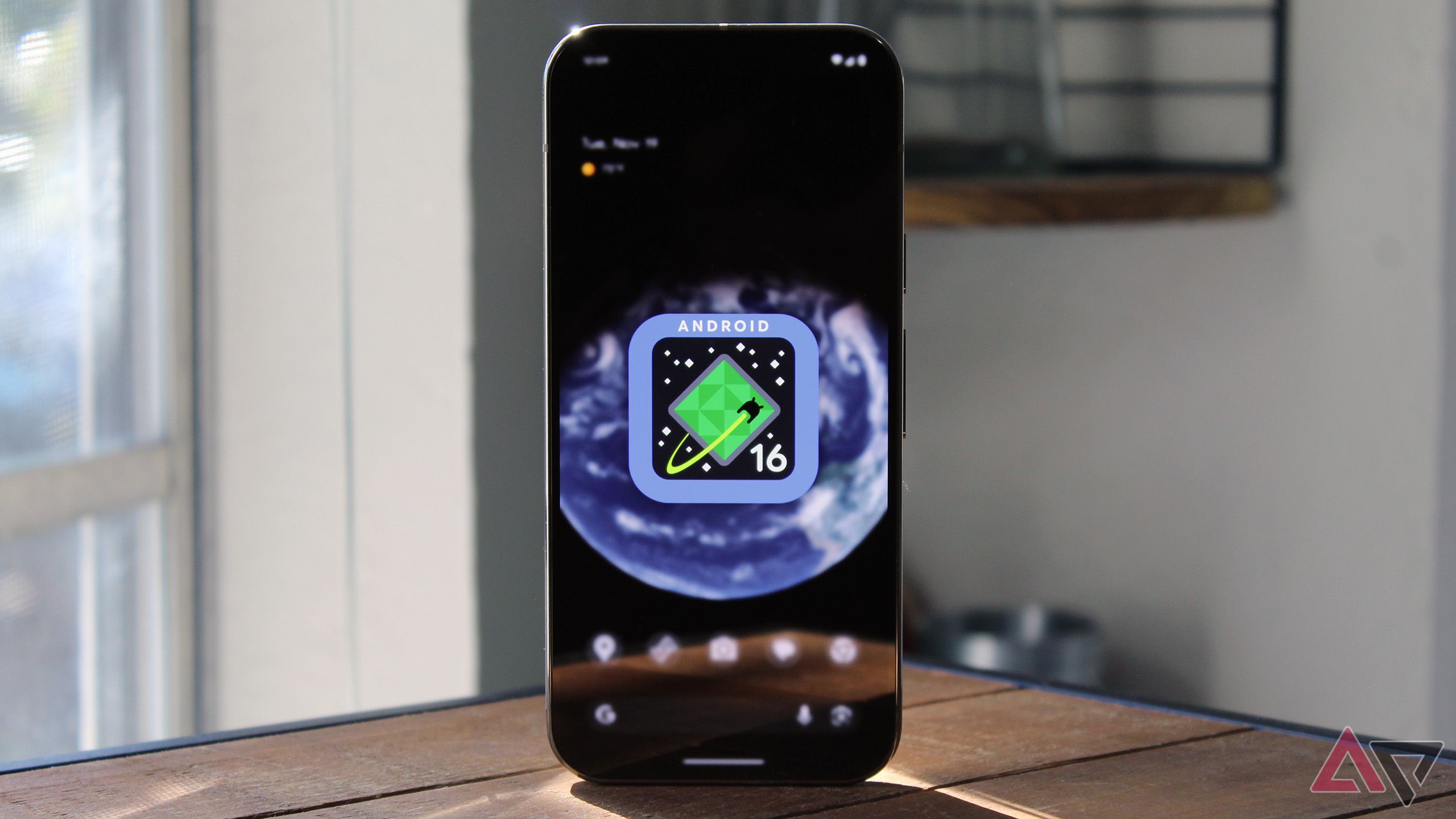 Image of the Android 16 logo on a Pixel 9 Pro's home screen with the phone sitting atop a wooden table.