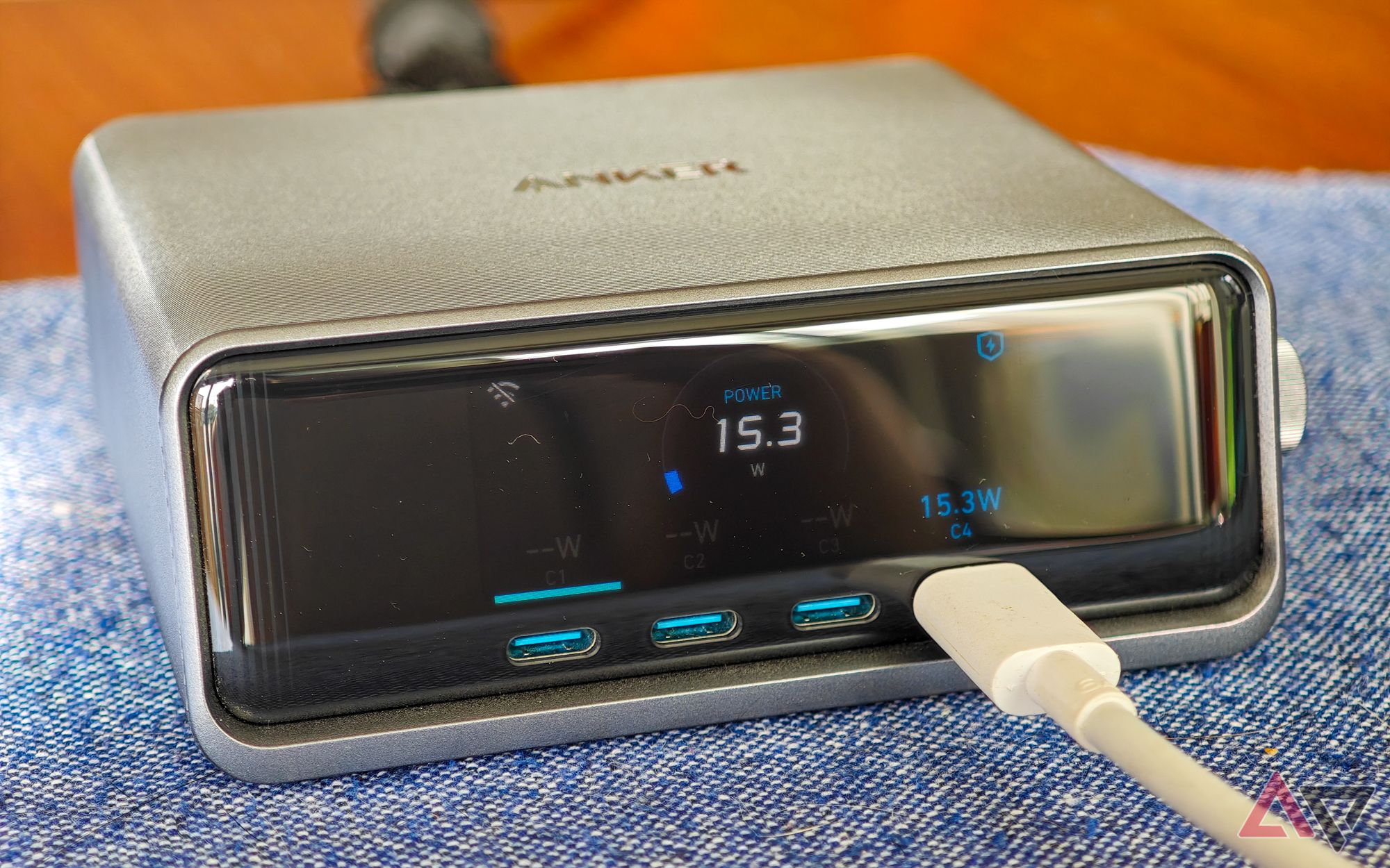 Anker Prime 250W desktop charger sitting on a blue cloth on a wooden table, with a white cable plugged into one USB-C port, showing 15.3W of power output