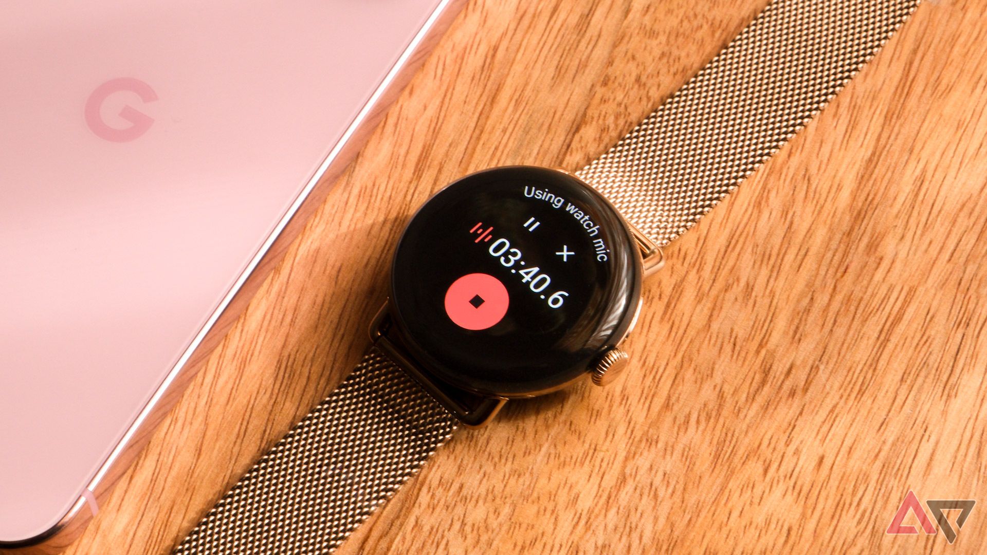 The Google Pixel Watch 3 with Recorder app on the display sits on a wood table next to a pink Google Pixel phone.