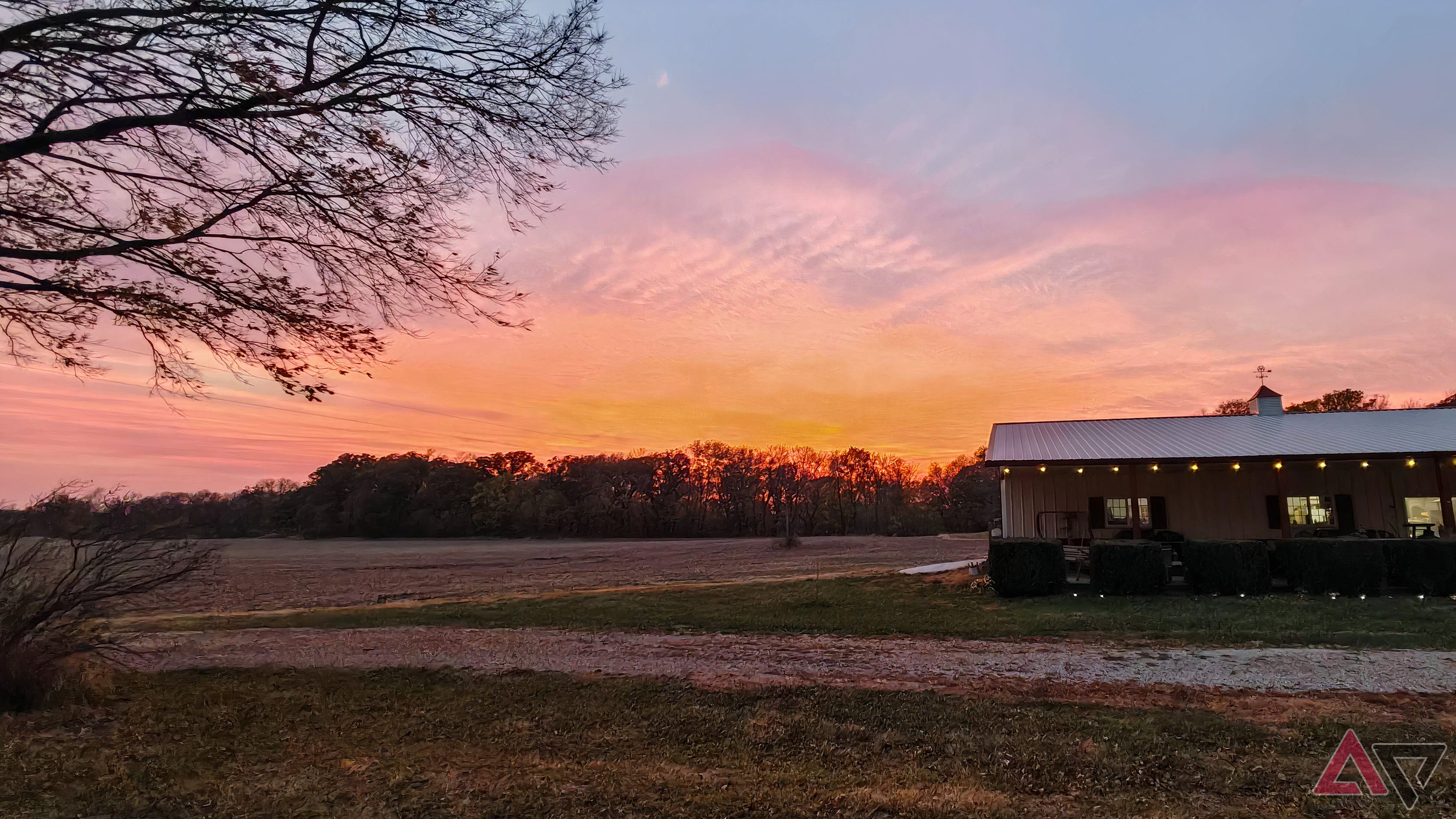 Colorful sunset over some trees and a field with a metal shop building in the foreground.