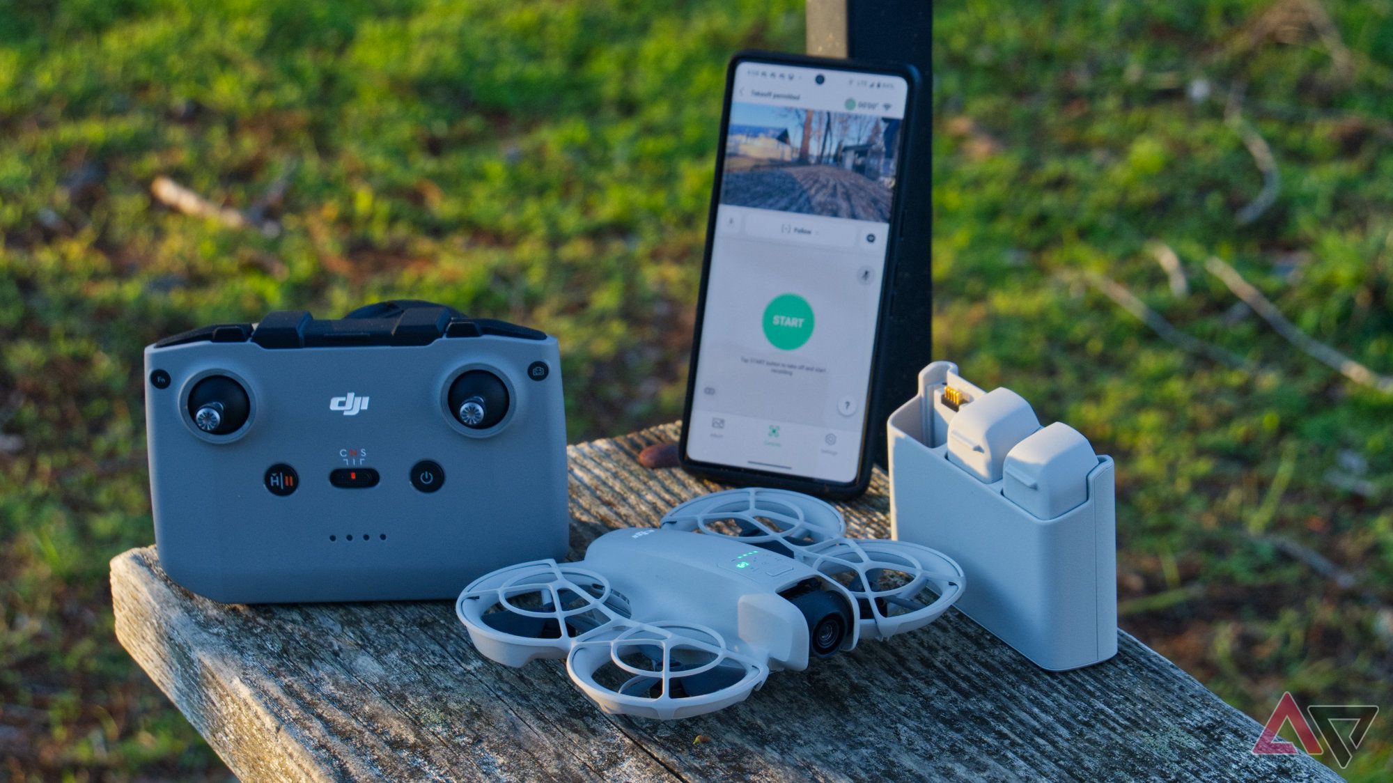 DJI Neo on a wooden bench next to the battery charger, RC-N3 controller, and a smartphone with the DJI Fly app showing a live view