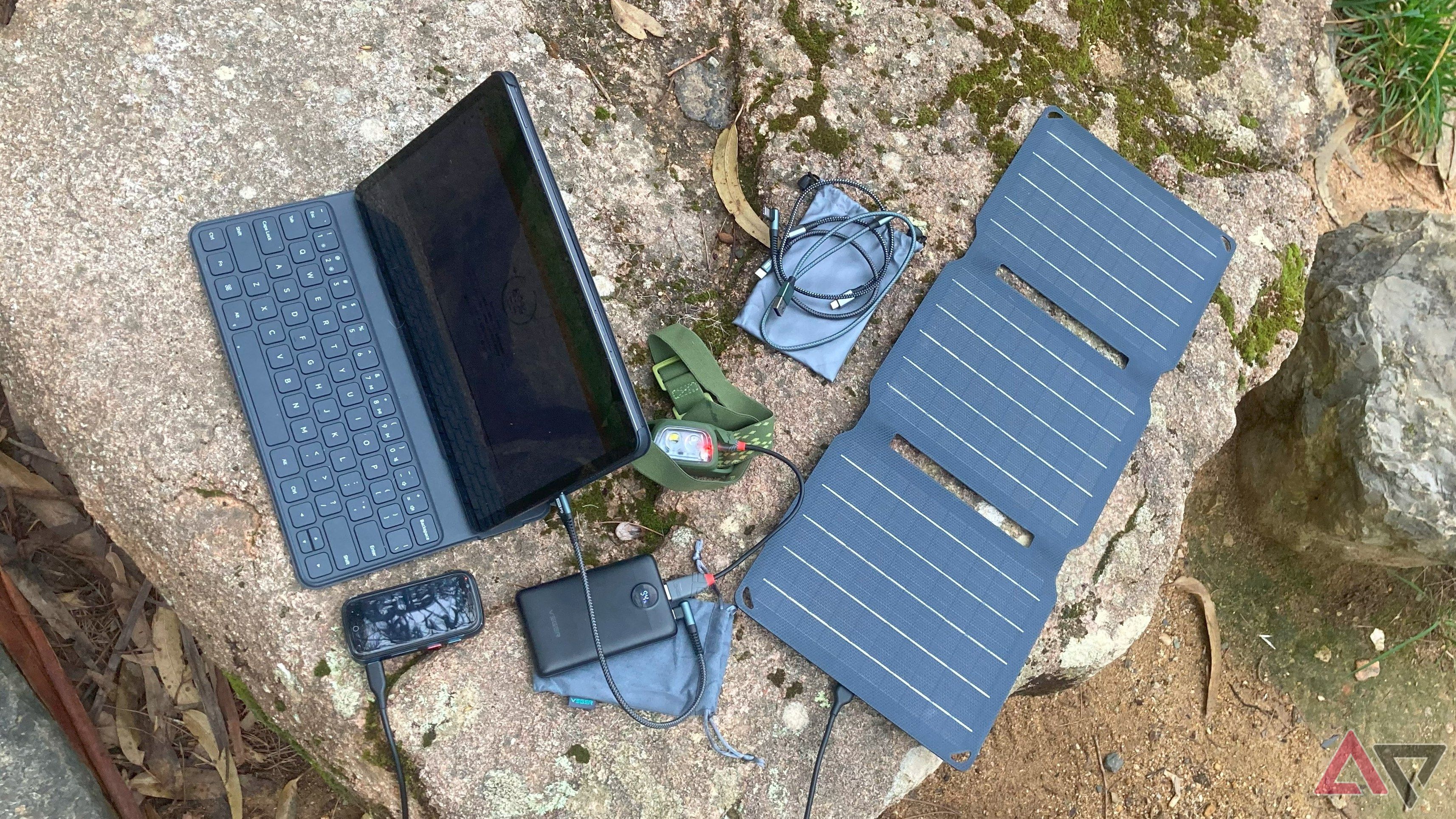 A charger, power bank, solar panel, smartphone, headlamp, and various cables on a rock table in nature