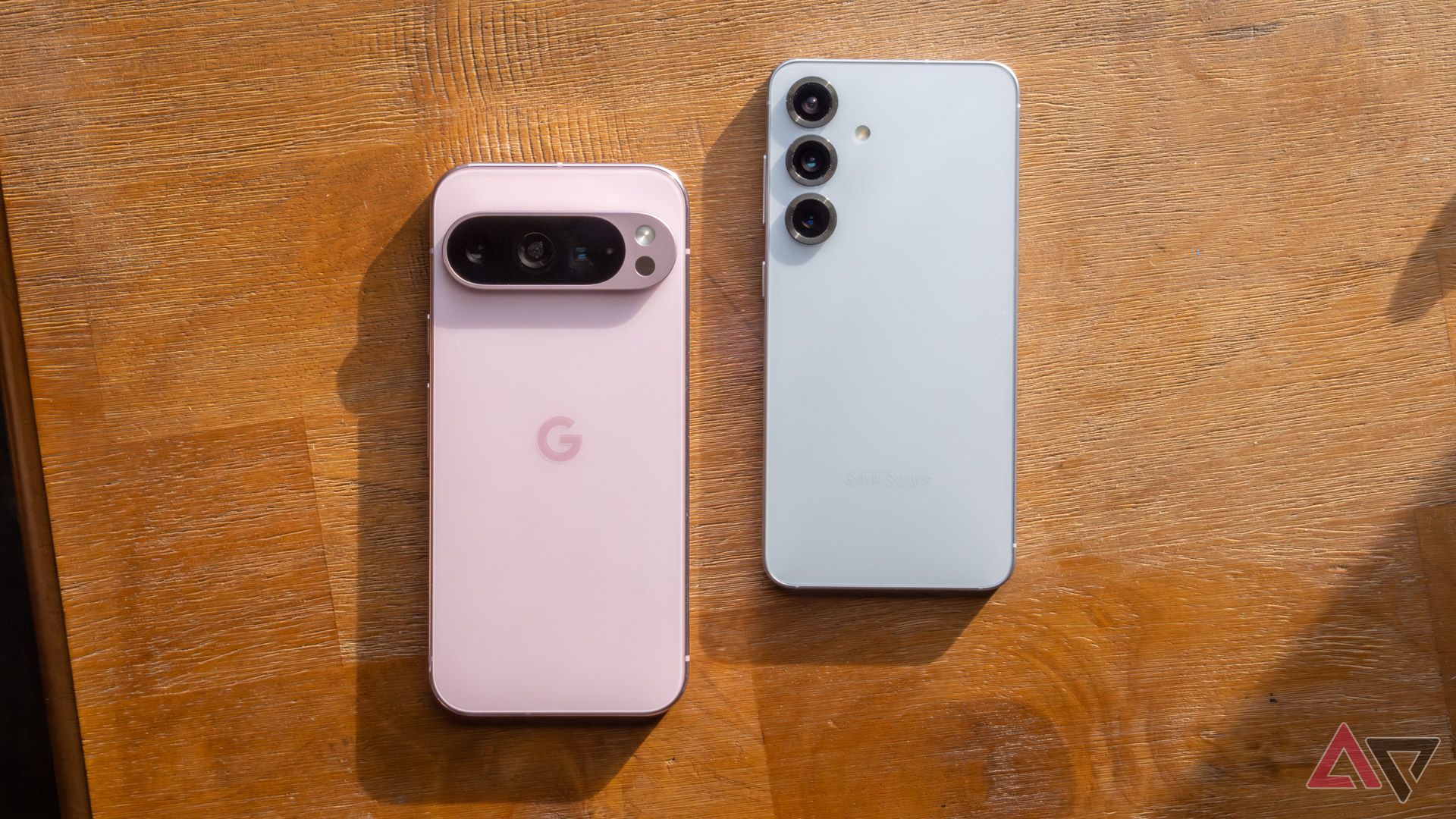 A pink Google Pixel 9 Pro next to a blue Samsung Galaxy S25, face down on a brown table